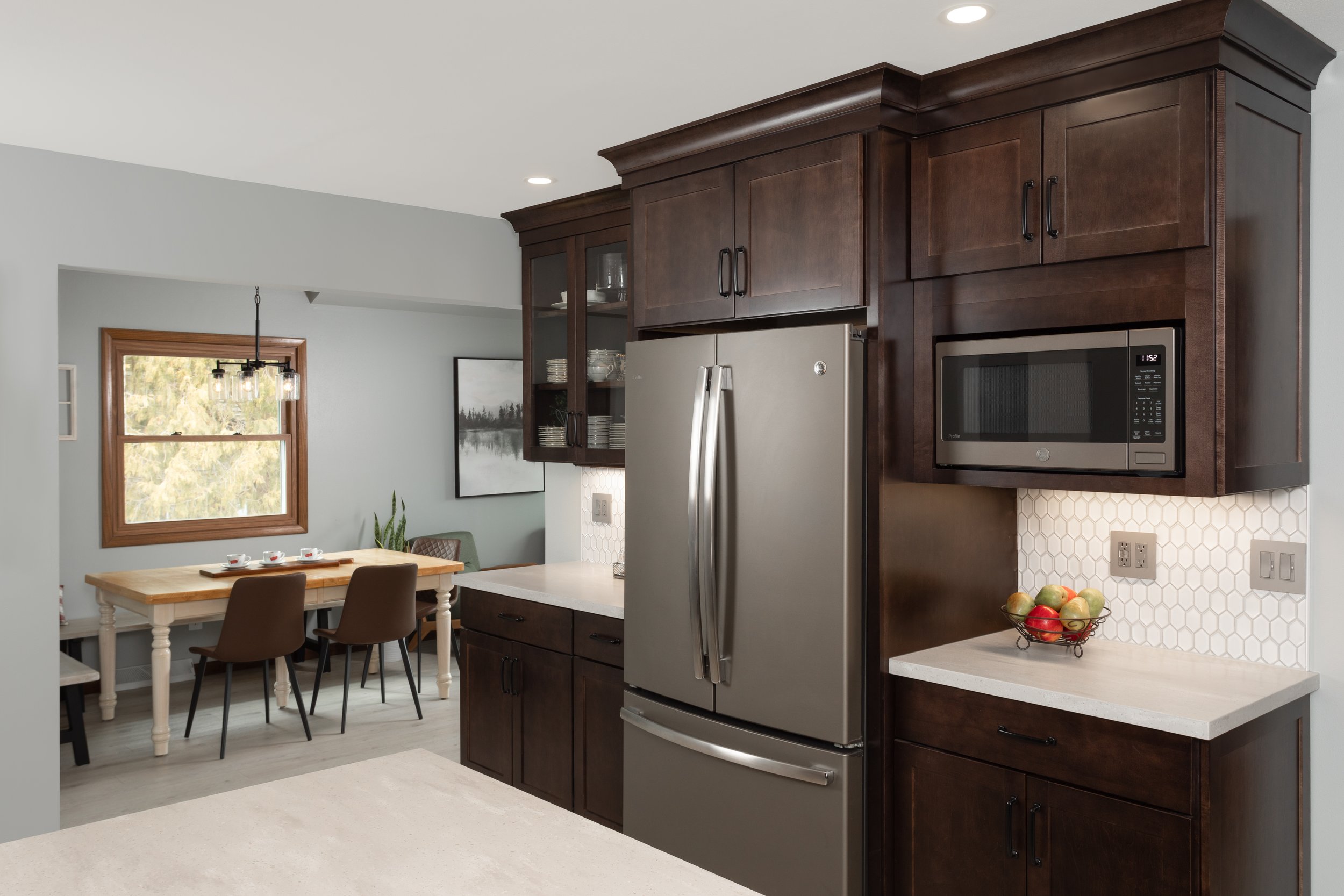 Kitchen with dark wood cabinets, stainless steel refrigerator, microwave, white hexagon tile backsplash, and fruit bowl on counter, view into dining area with window, table, and chairs