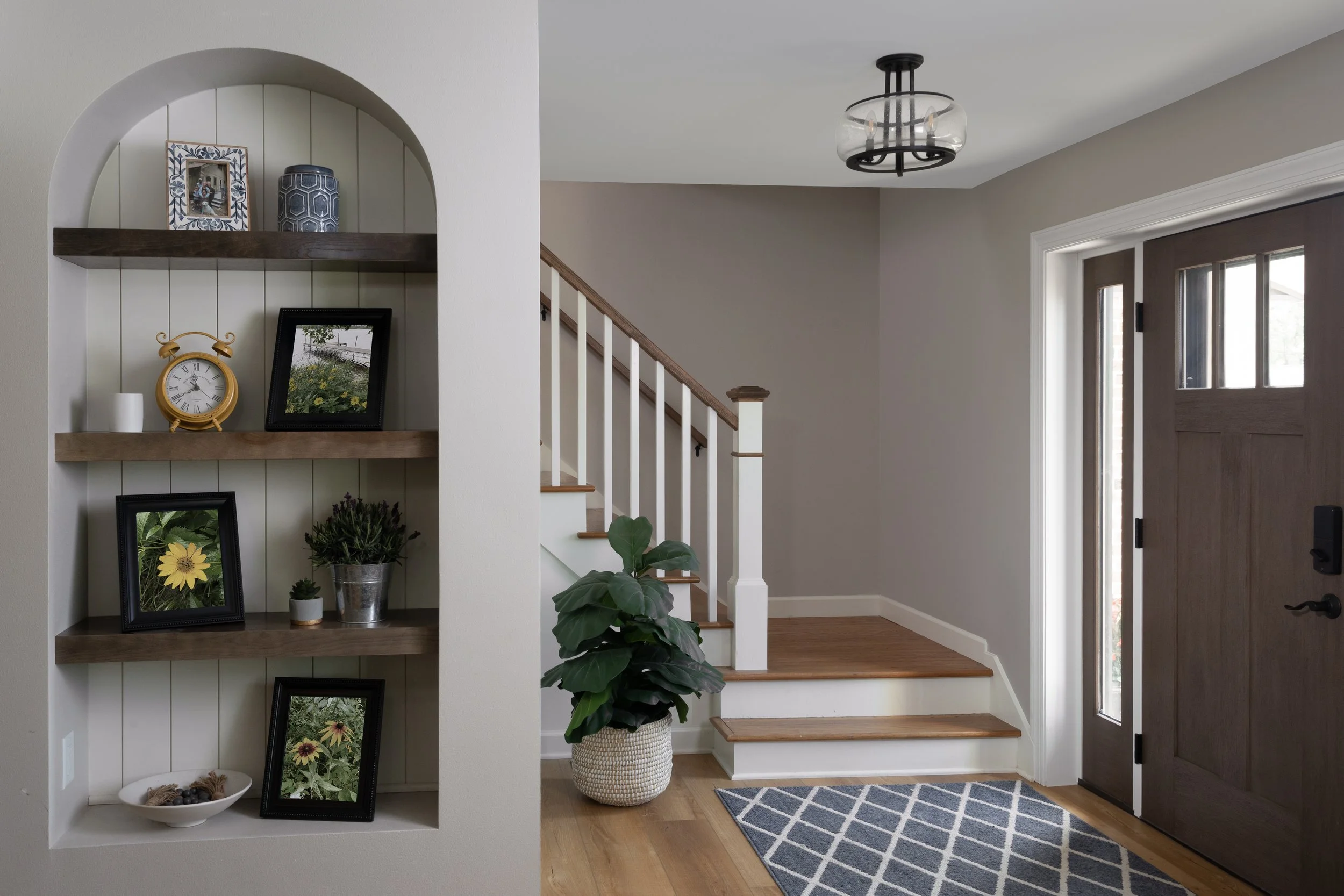 Entryway with a dark wooden front door, a stairway with wooden steps and white railings, a patterned blue and white rug, a potted plant in a white woven basket, and a built-in shelf with picture frames, a yellow alarm clock, and decorative items.
