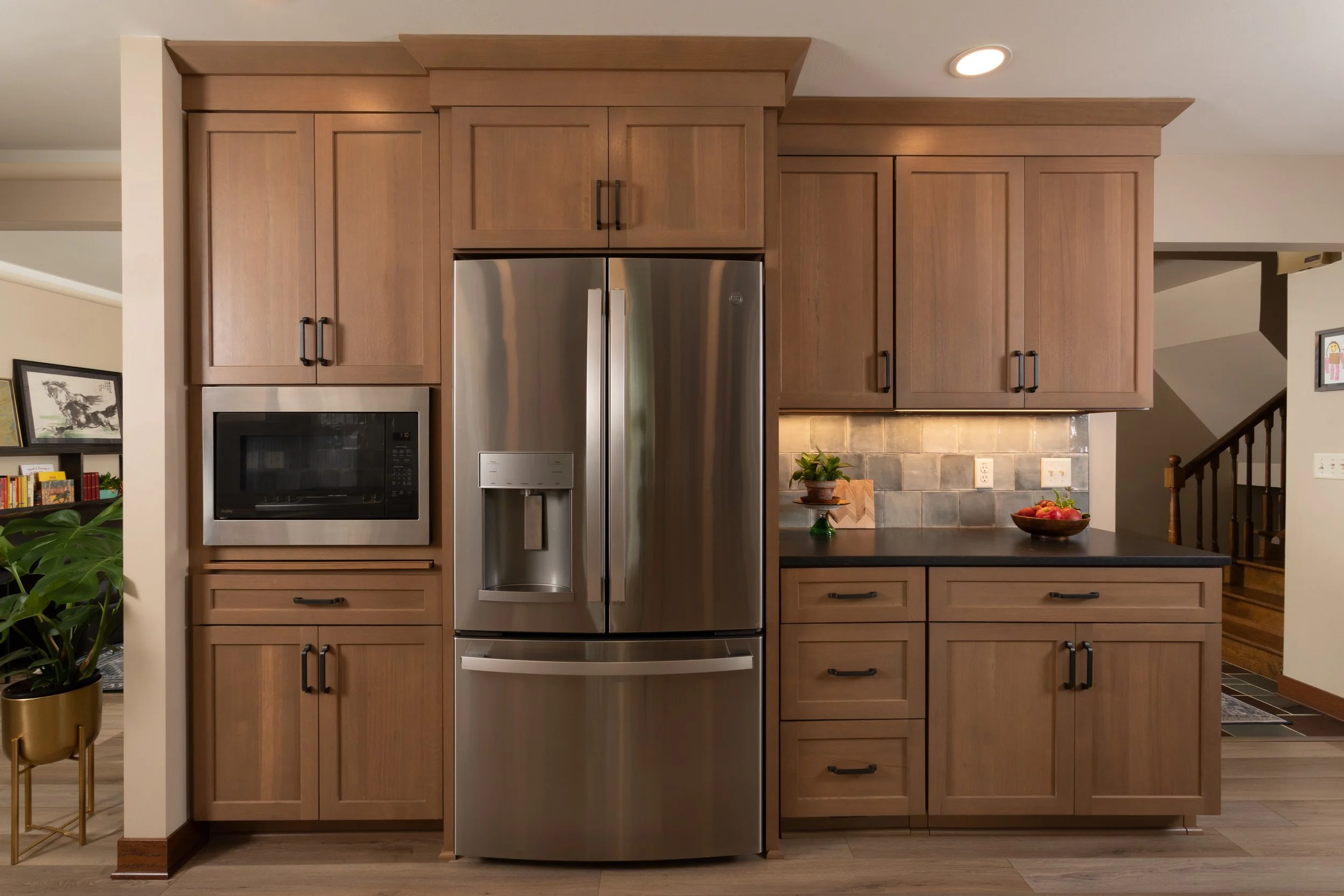 Kitchen with wooden cabinets, stainless steel refrigerator, built-in microwave, black countertop, decorative plants, and a bowl of apples.