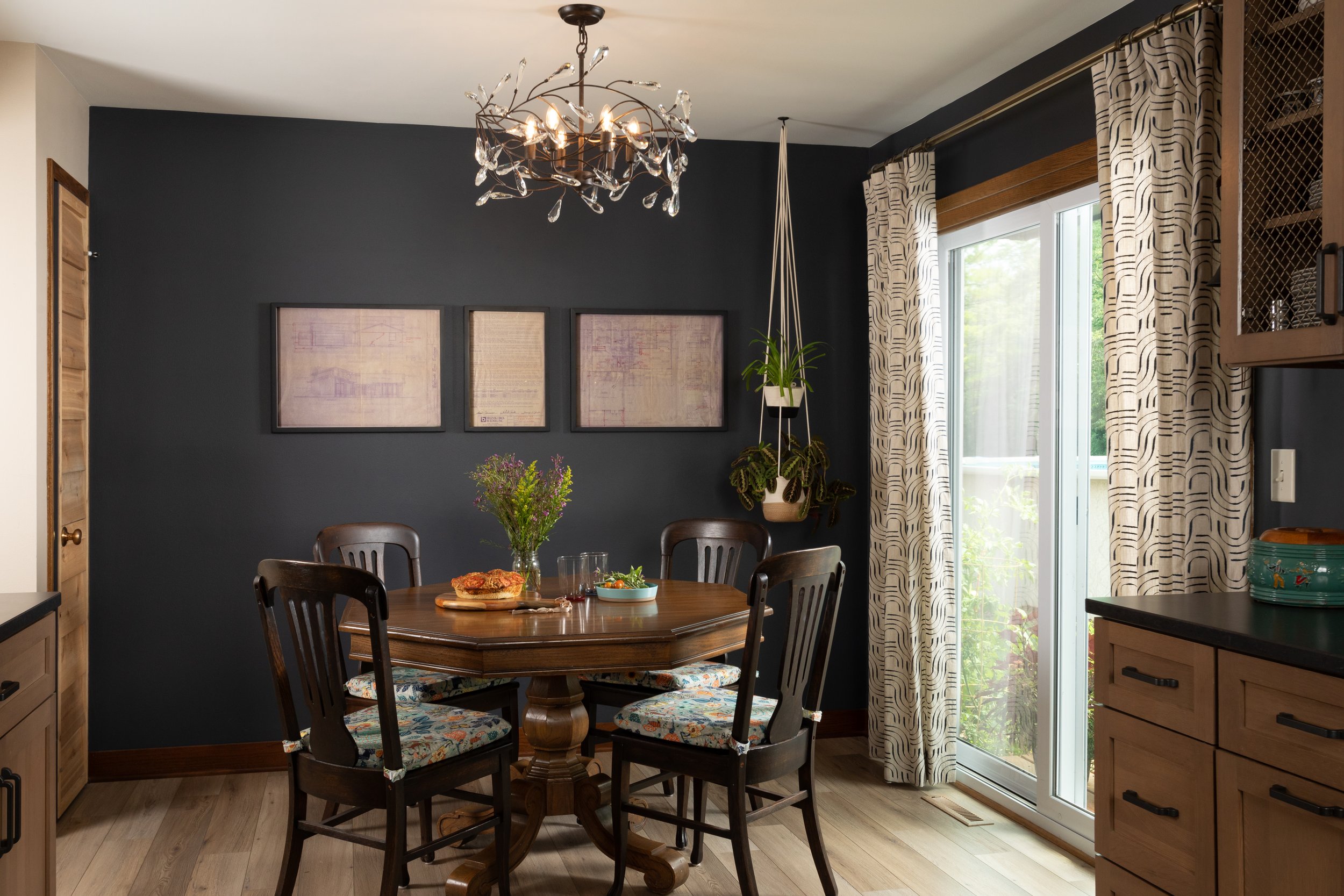 Dining room with dark accent wall, round wooden table set with food and flowers, four chairs with patterned cushions, sliding glass door with patterned curtains, hanging plant, and ceiling chandelier with crystal accents.
