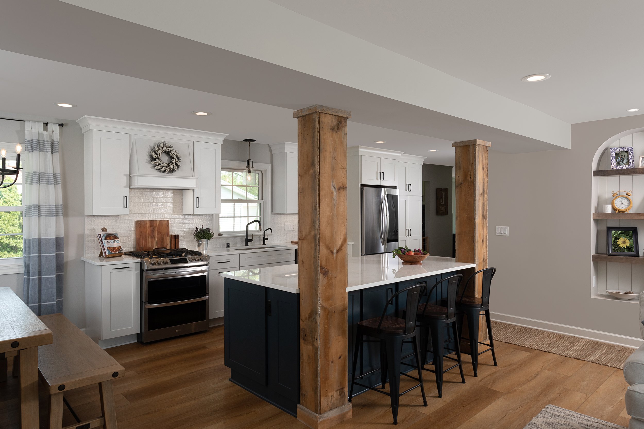 Modern kitchen with white cabinets, stainless steel appliances, a kitchen island with a dark base, and wooden support columns. There's a window above the sink, a bowl of fruit on the island, and decorative shelves on the wall.