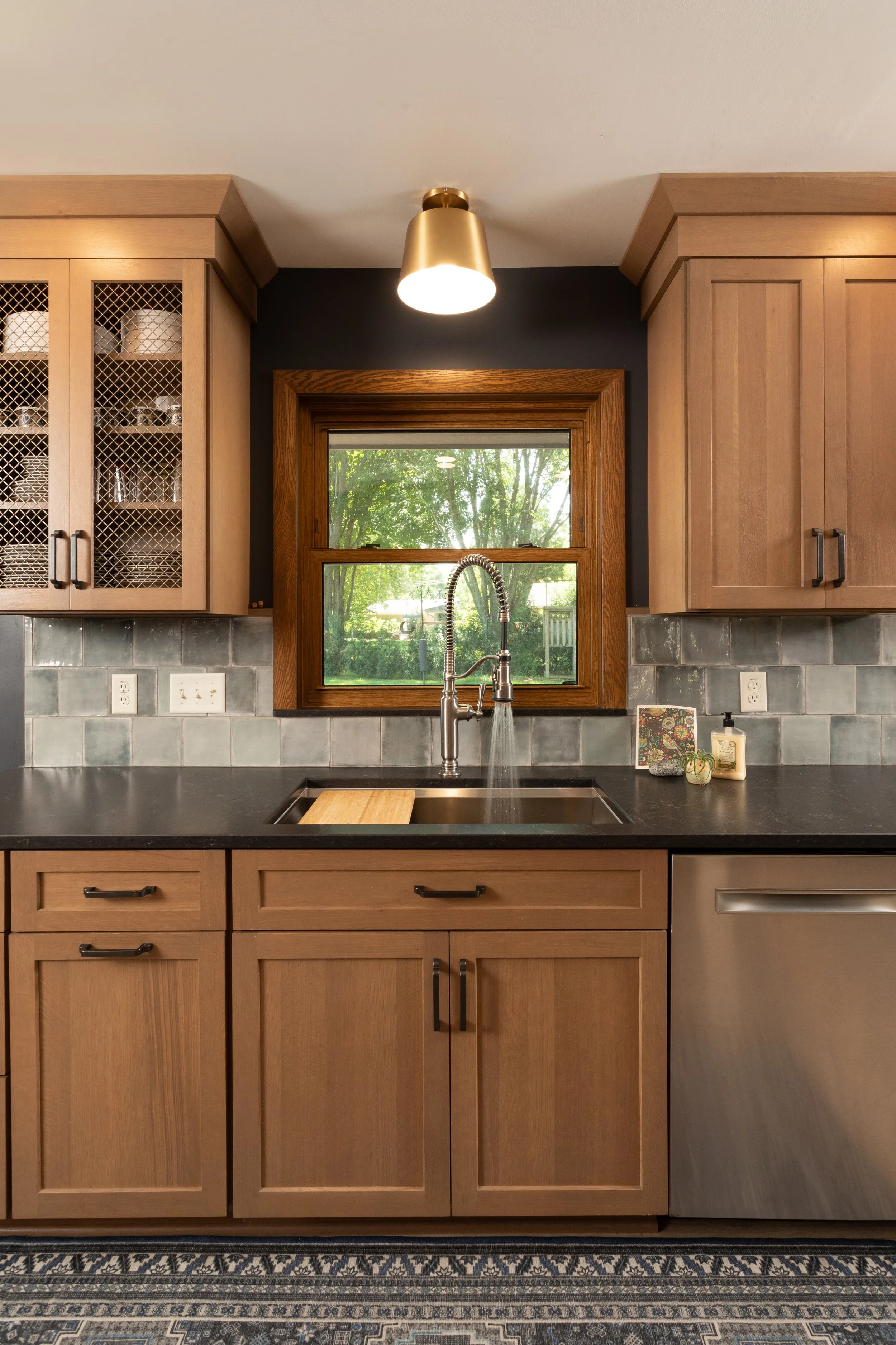 Kitchen with wooden cabinets, a black countertop, a stainless steel sink under a window showing trees outside, and a modern light fixture on the ceiling.