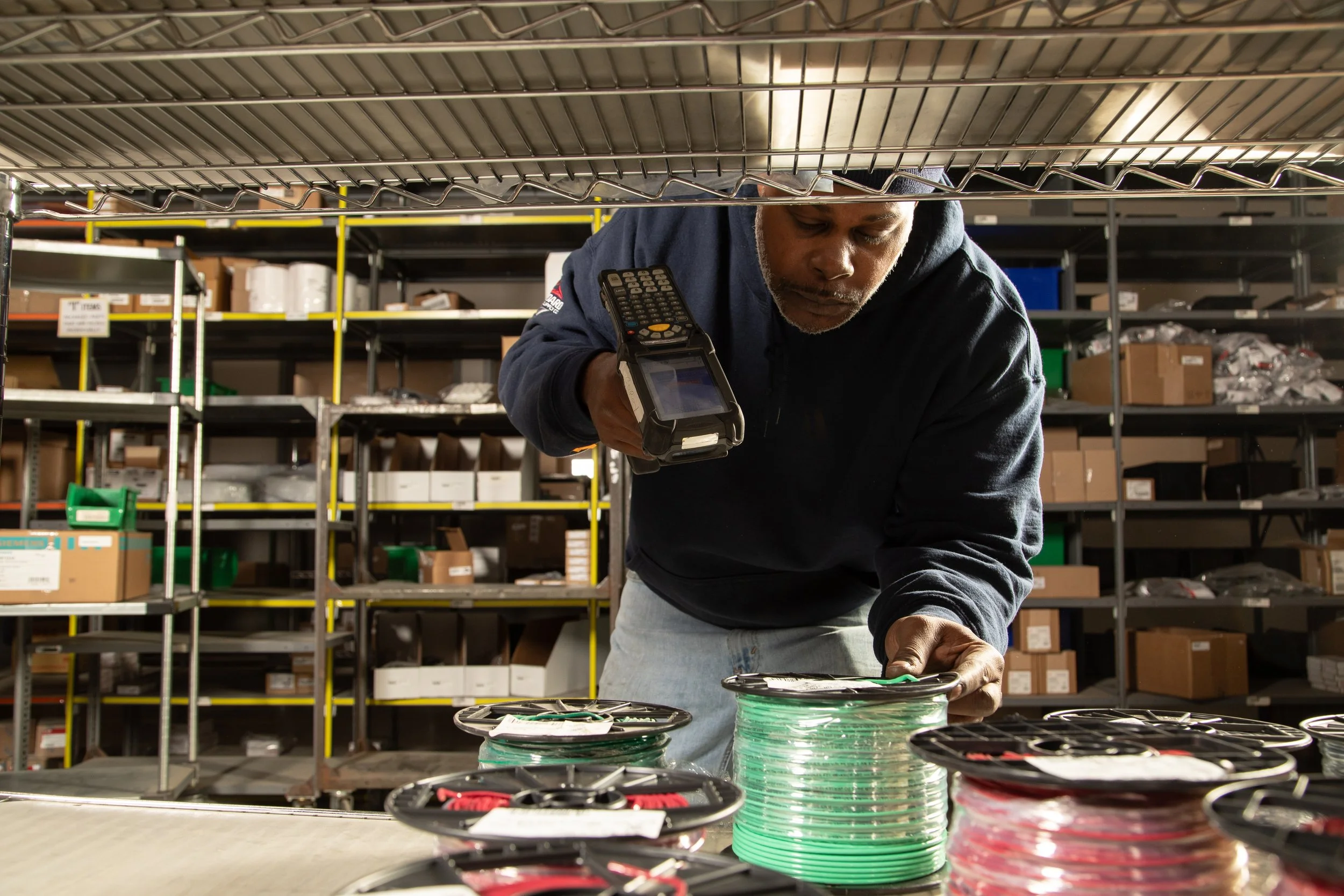 A man working in a warehouse, examining wires or cables on a table with shelves filled with boxes and supplies in the background.