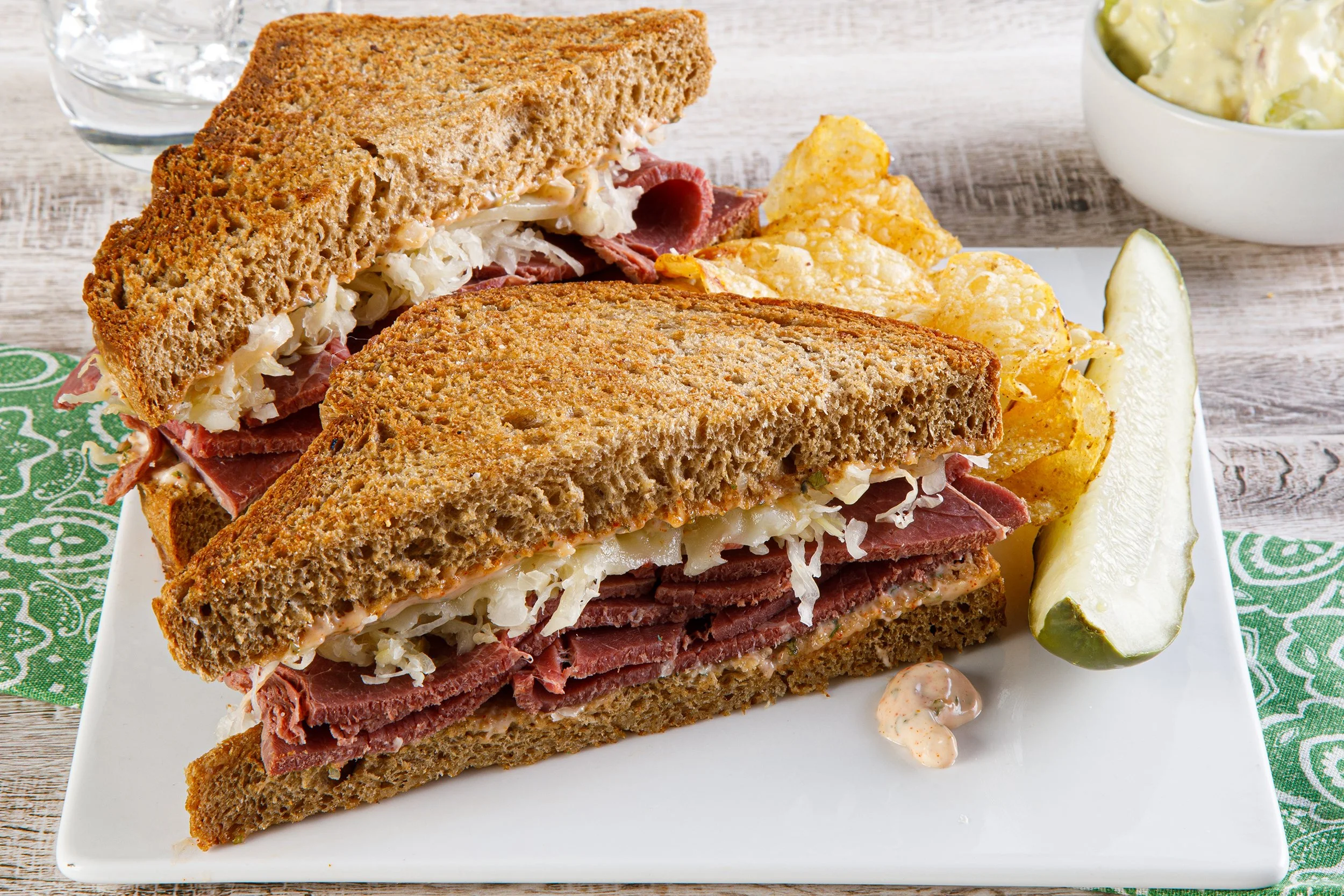 Food photography by Tom Krueger, photographer, Milwaukee. Sandwich with rye bread filled with sliced roast beef, coleslaw, and cheese, served with potato chips, a lemon wedge, and a small dollop of sauce on a white plate, on a green patterned napkin.