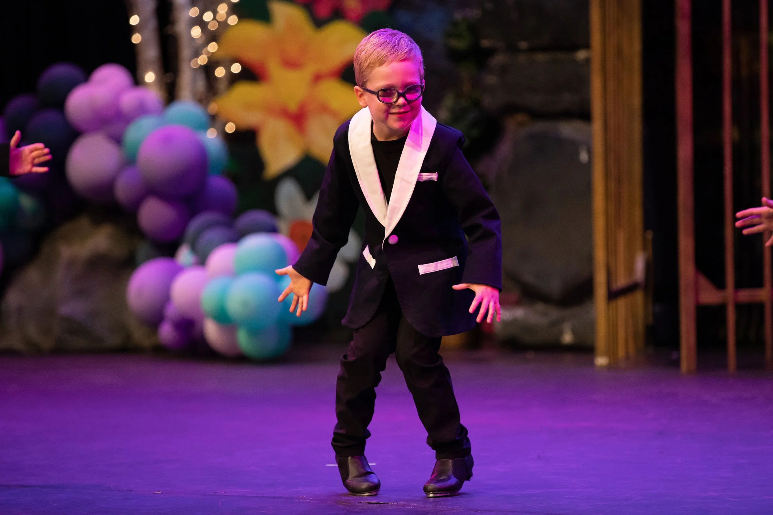 A young boy with glasses and a tuxedo, dancing on stage with colorful balloons and decorations in the background.
