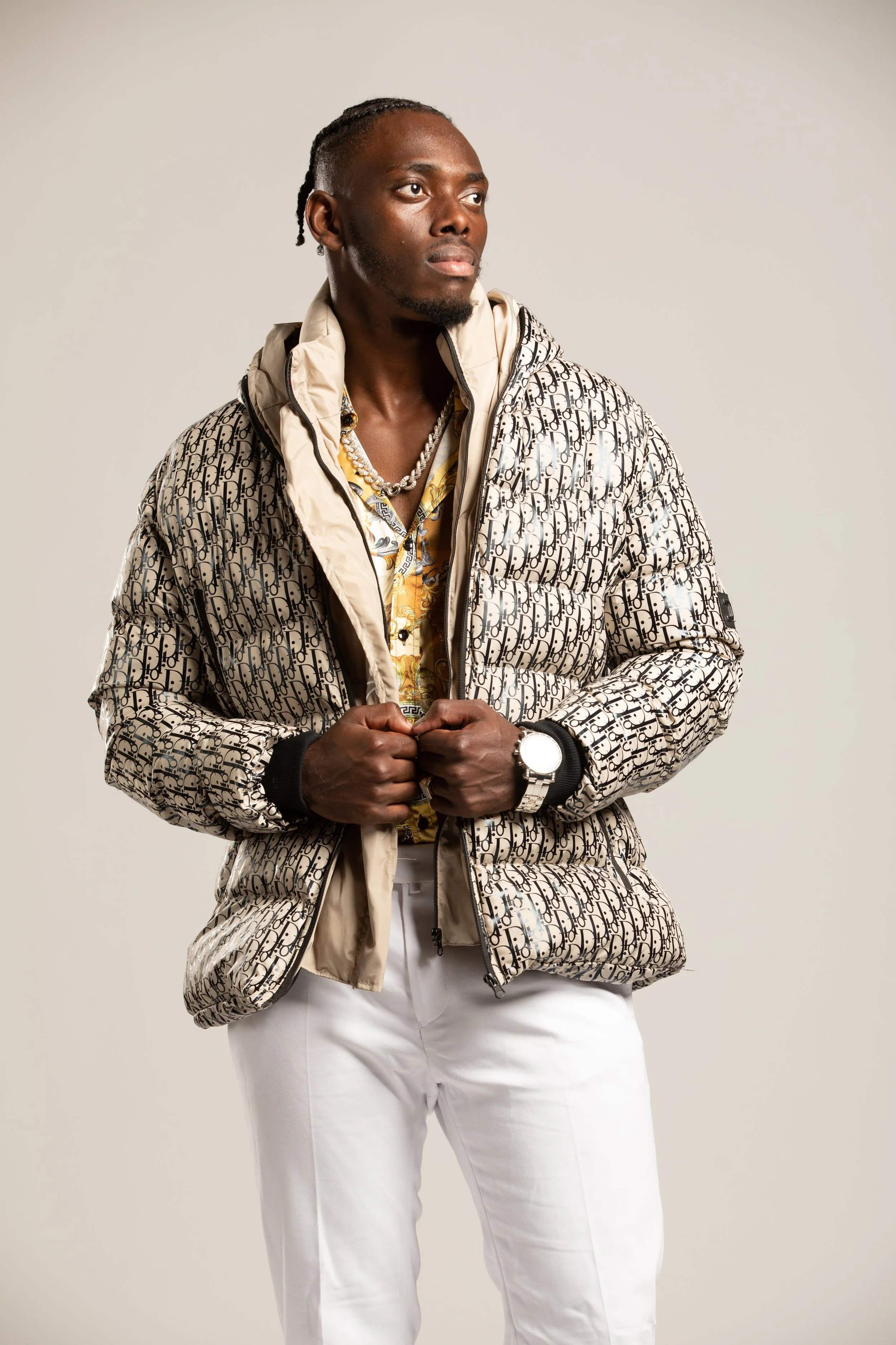 A man with dark skin and braids looking to his left, wearing a designer patterned jacket, a watch, a necklace, and white pants posed against a plain light background. Photo by studio portrait photographer Tom Krueger, Milwaukee, Wisconsin.