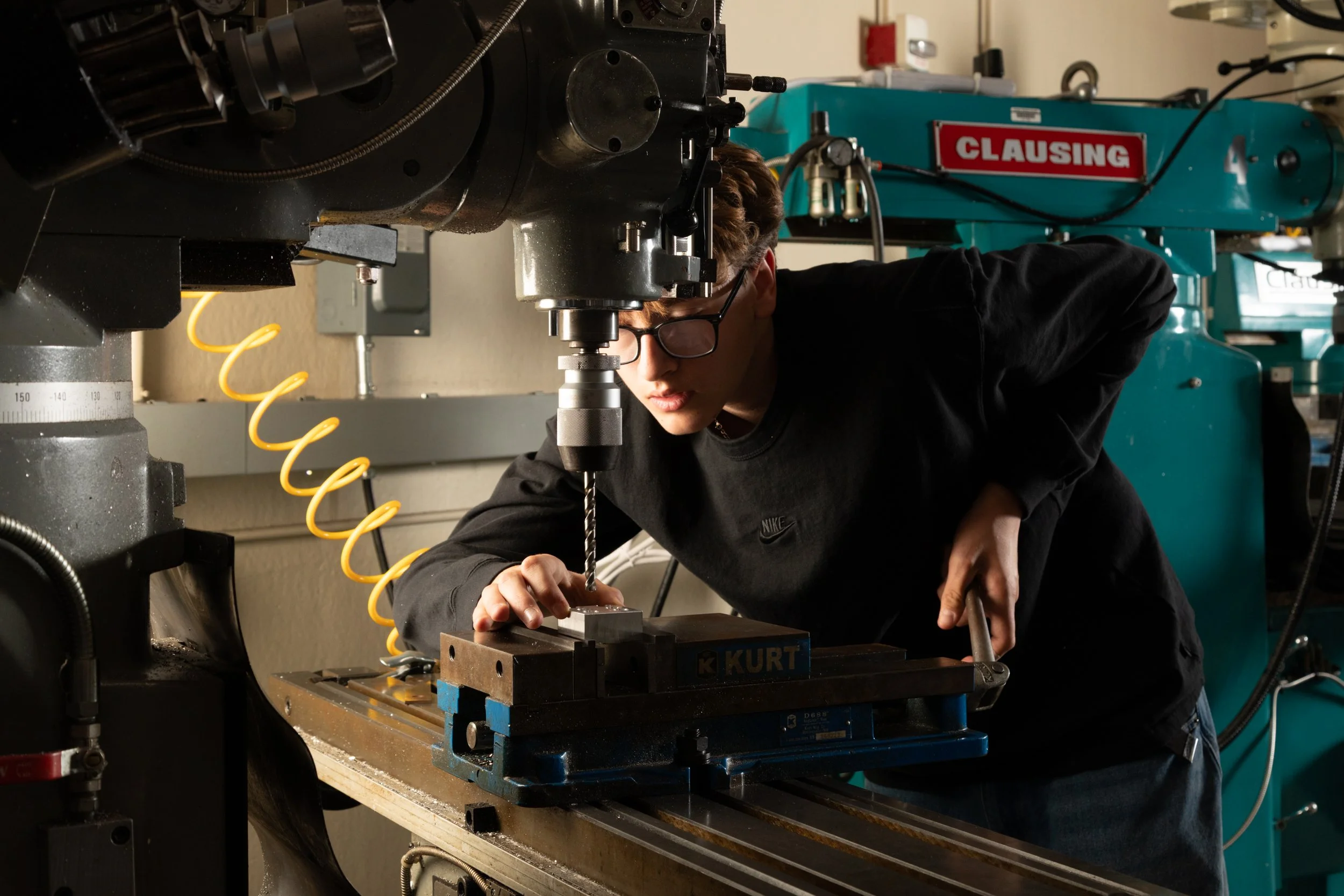 A young man with glasses is operating a milling machine in a workshop. He is focused on the metal piece he is working on, and there are various mechanical and electronic equipment around him.