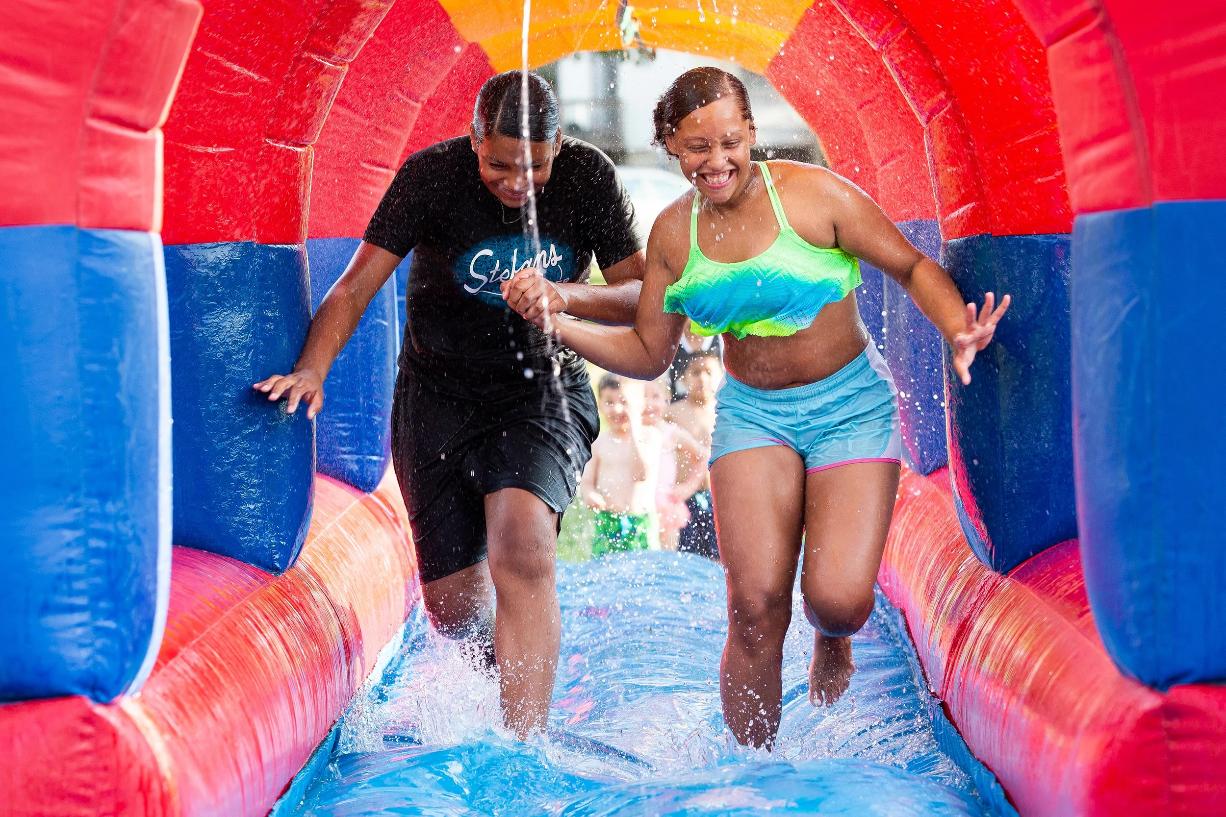 Two women are smiling and having fun as they walk through a colorful inflatable water slide, holding hands. Water is splashing around them and they are wearing swimwear.