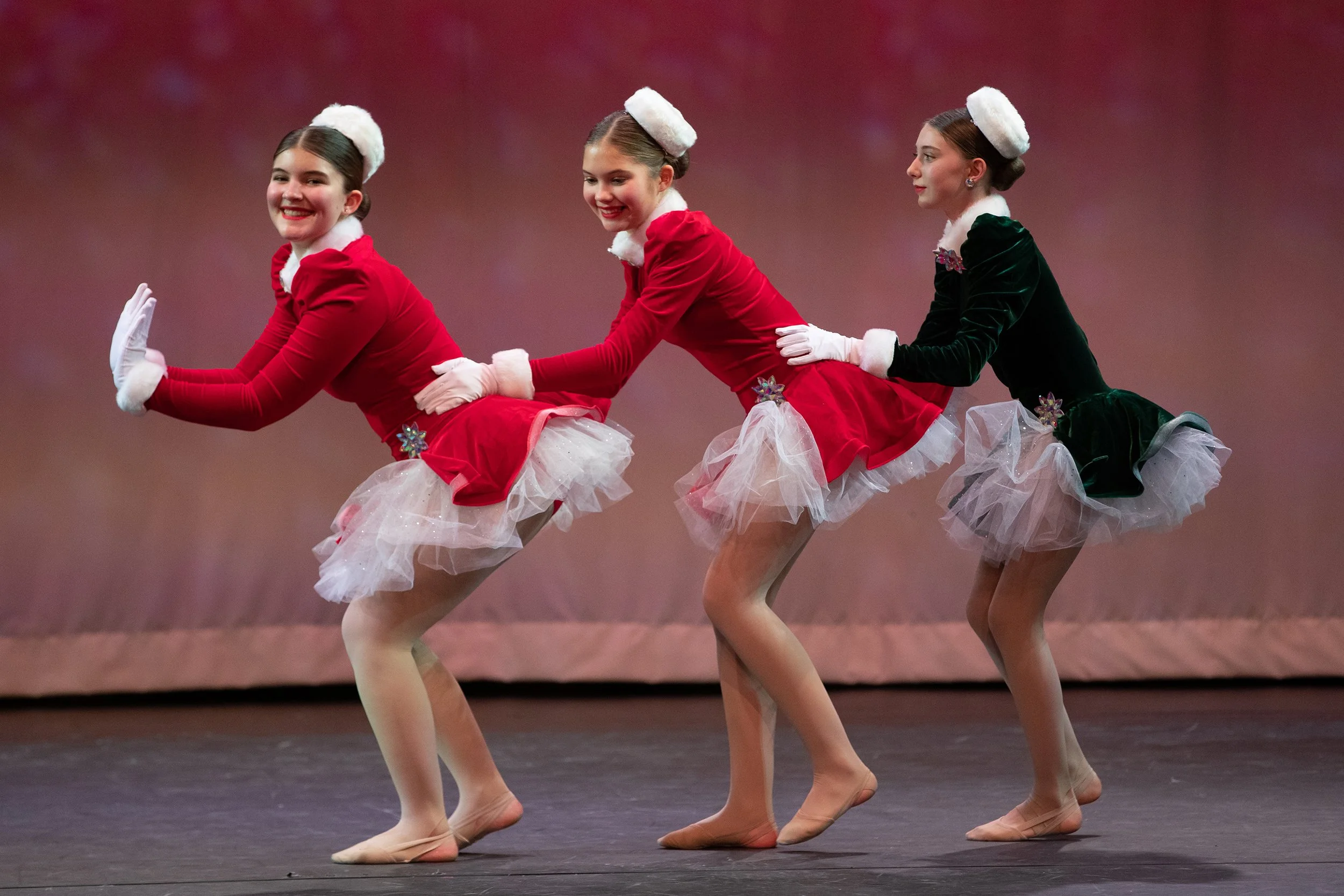 Three young girls in Christmas-themed costumes performing a dance on stage, each wearing a velvet dress with a tutu, tights, gloves, and a fluffy headpiece.