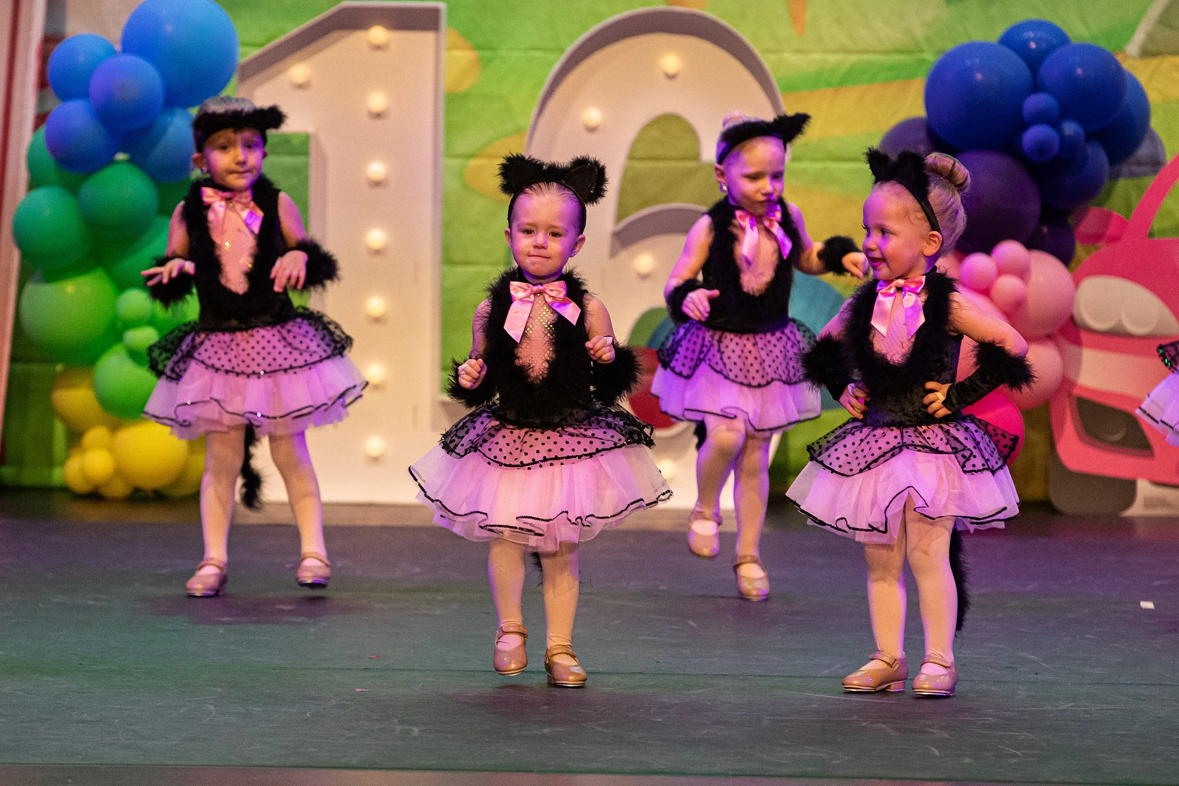 Young girls dressed in pink and black cat costumes perform a dance on a decorated stage at a children's party or event.