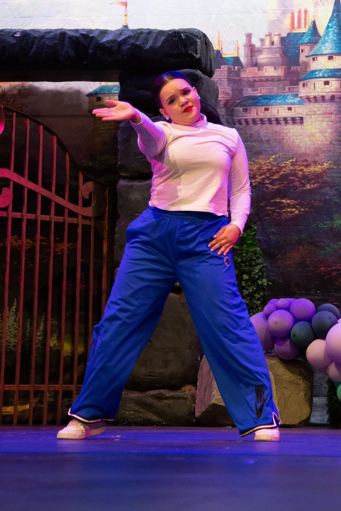 Performer in costume on stage with castle backdrop, wearing blue pants and white top, striking a dramatic pose.