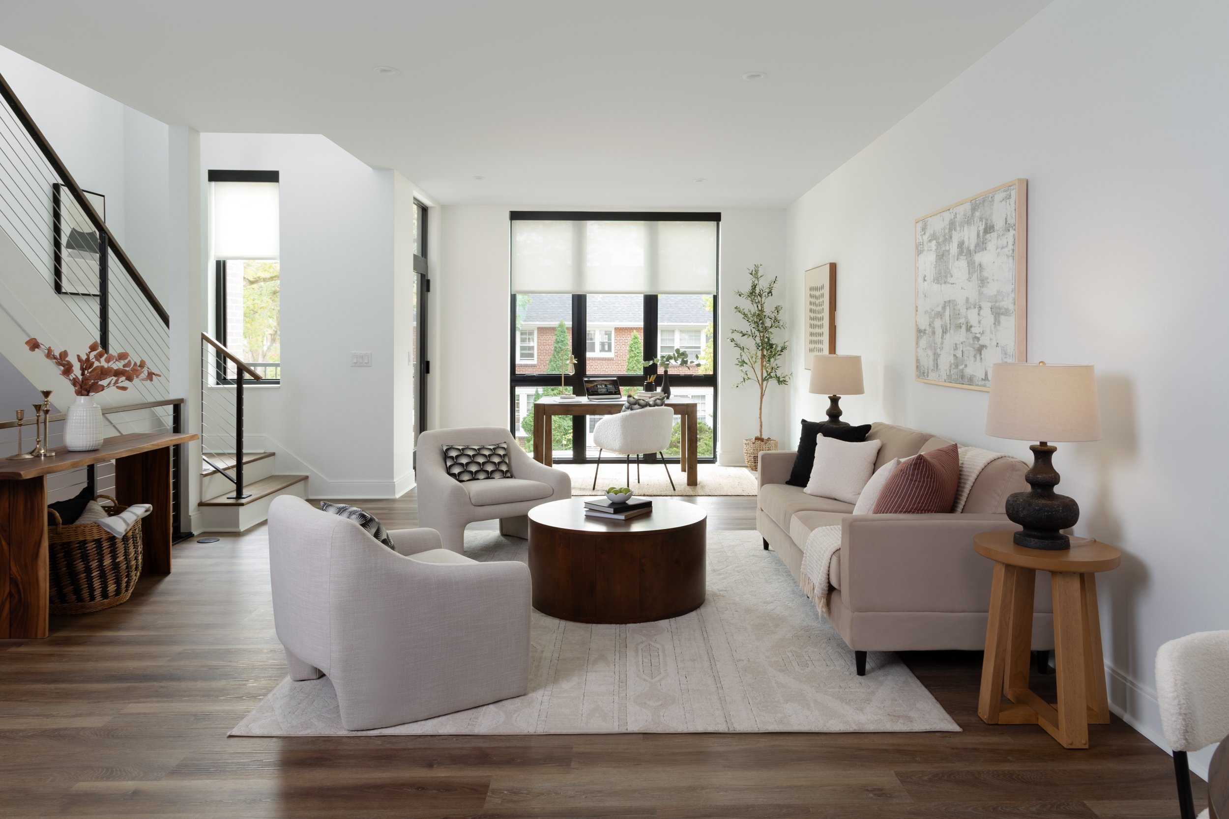 Bright living room with white walls, hardwood floors, beige sofa, two white armchairs, a round wooden coffee table, and a small desk near large windows with white blinds, decorated with plants and artworks.