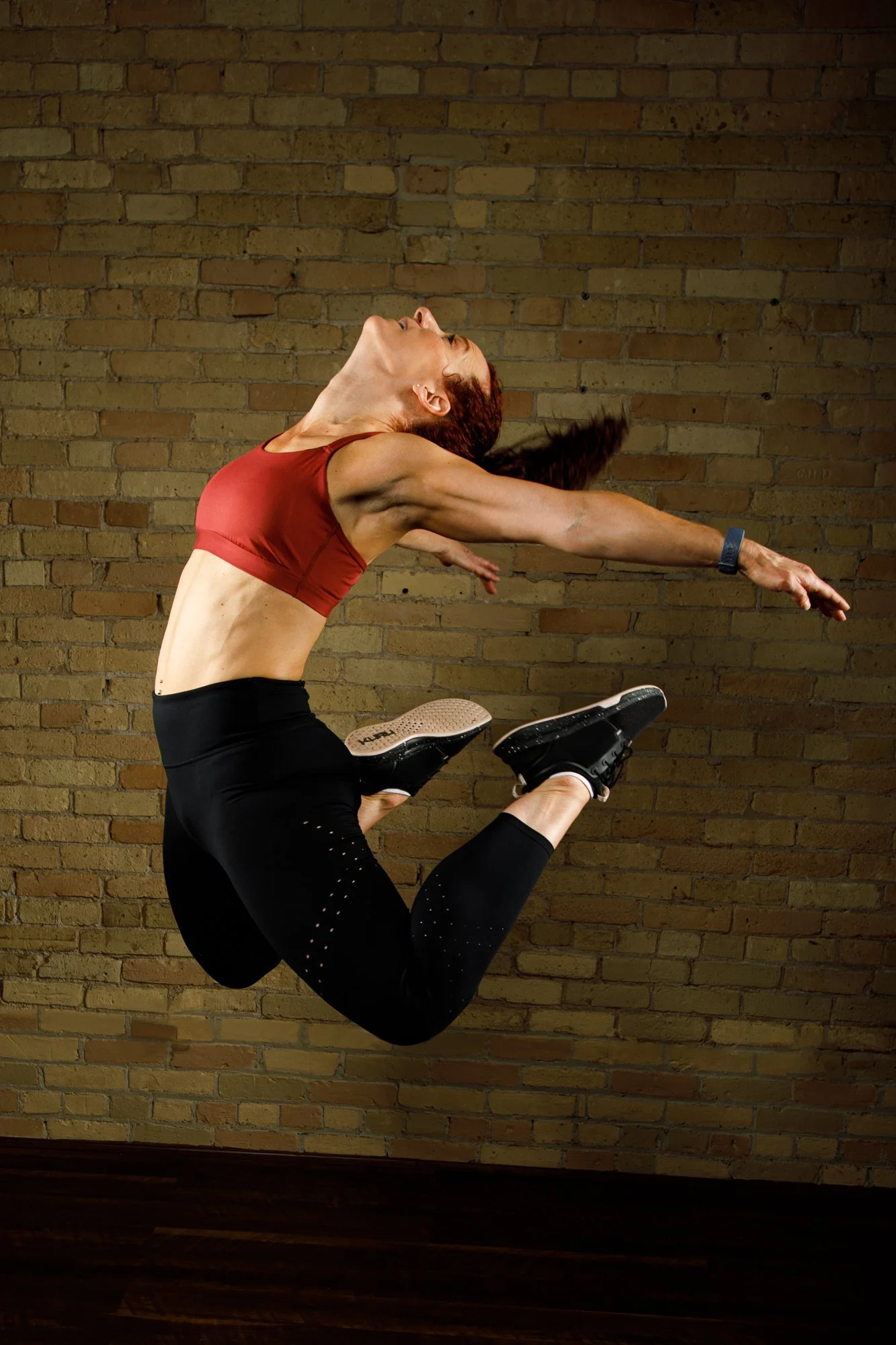 A woman in a red sports bra and black leggings is jumping in front of a brick wall, holding her legs with her arms extended backward and her head tilted back.