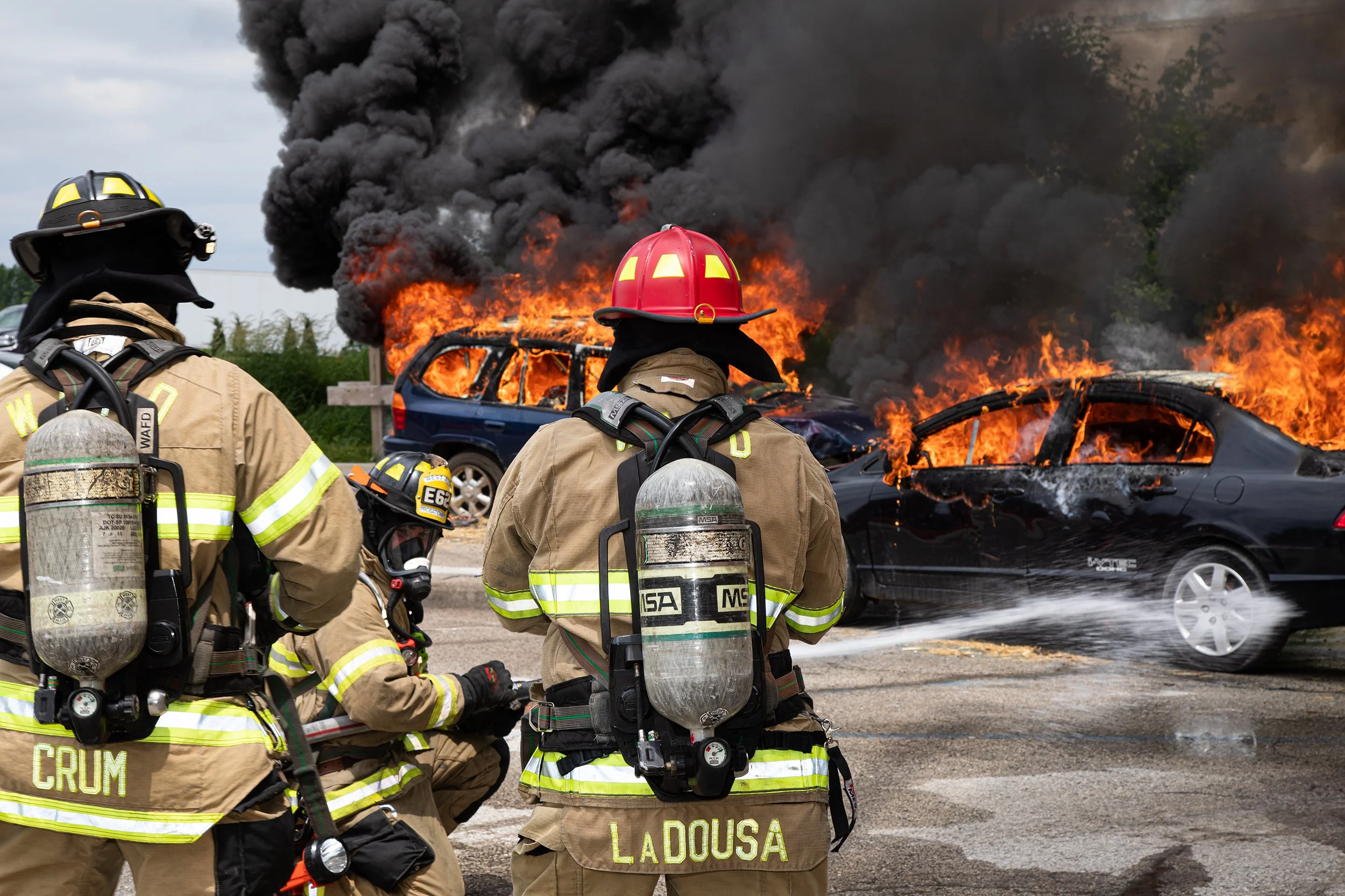 Firefighters in protective gear and helmets stand in front of a car engulfed in flames with thick black smoke rising into the sky.