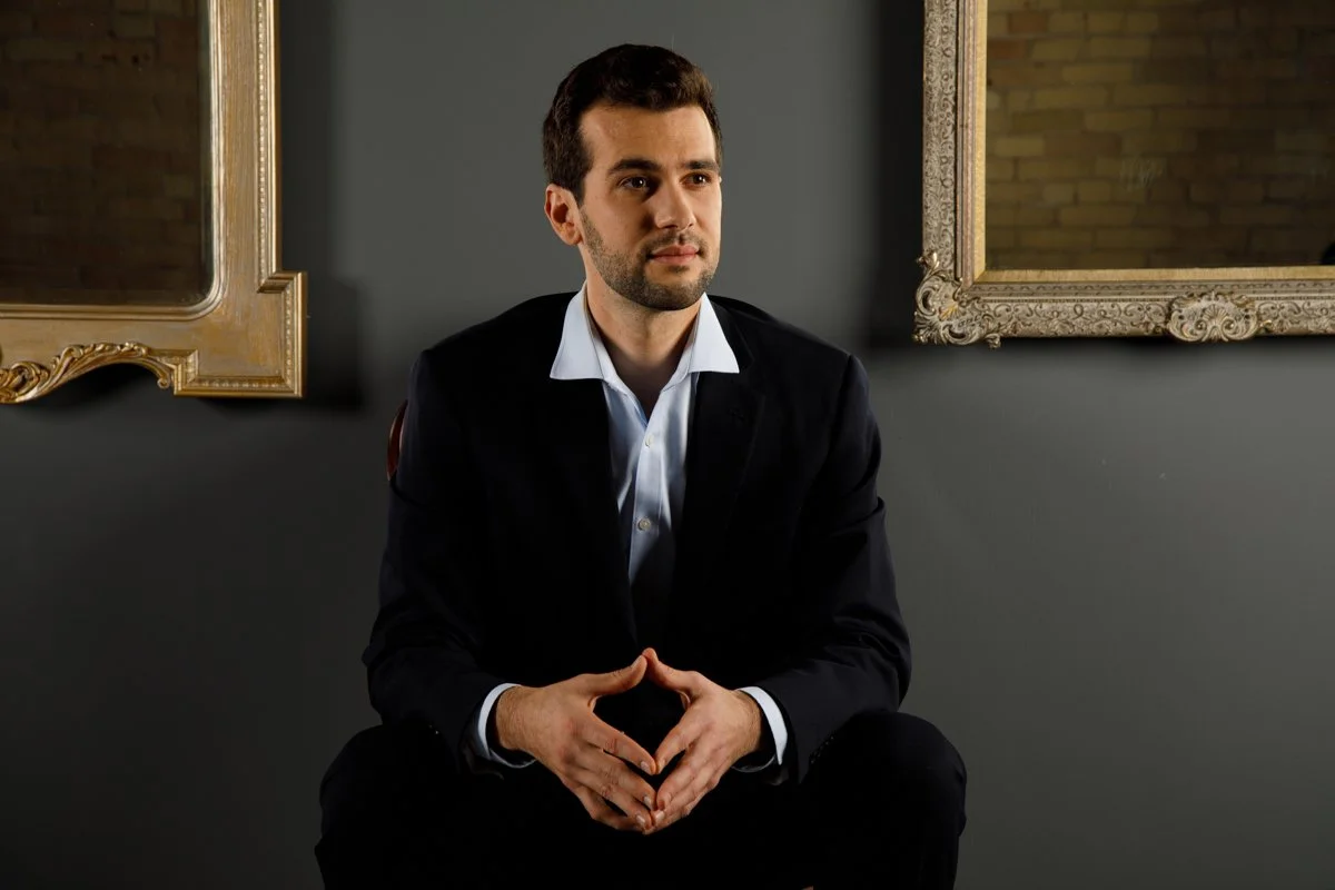 A man in a black suit and white shirt sitting in front of a dark gray wall with two ornate gold-framed mirrors.