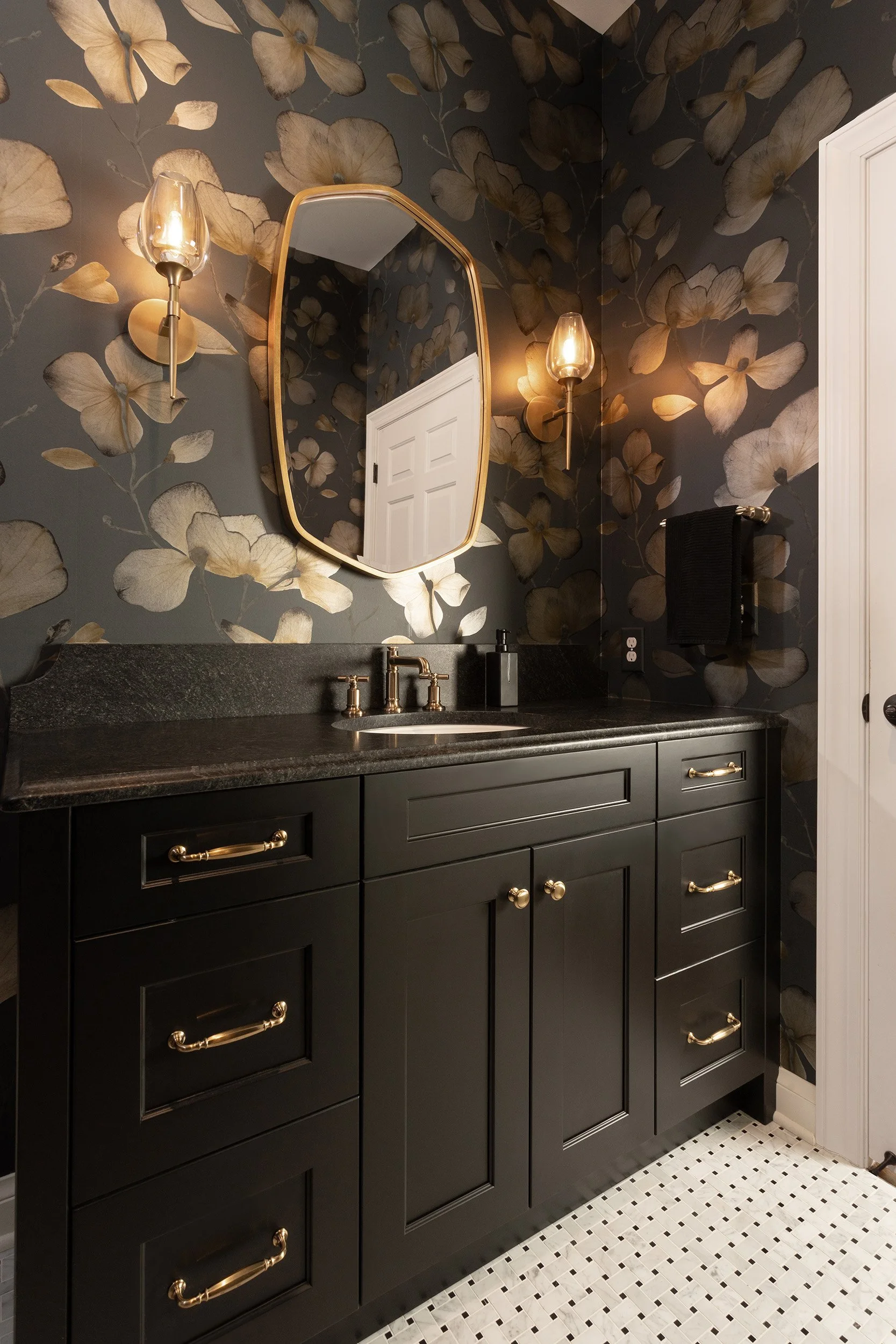 A bathroom vanity with black cabinetry, a black countertop, and gold hardware. Two wall-mounted sconces with gold bases and glass shades, a mirror with a gold frame, a faucet, soap dispenser, and black towel on the wall. Floral wallpaper with large b