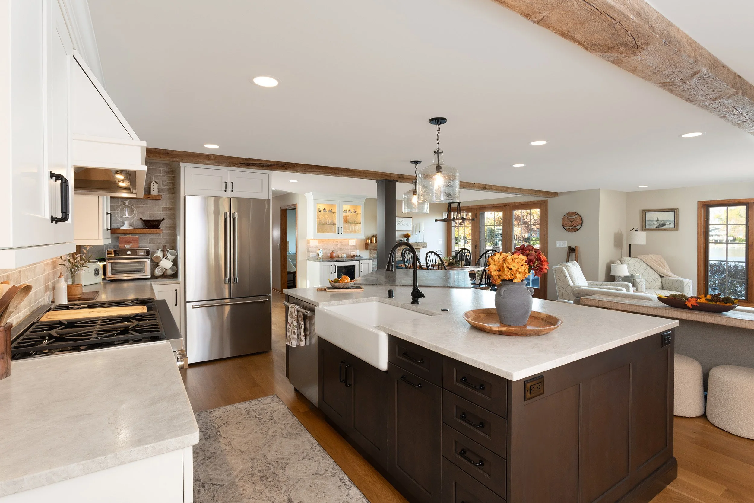 Open-concept kitchen and living area with white cabinets, stainless steel refrigerator, island with sink, and a sitting area with large windows, wooden accents, and colorful flower arrangements.