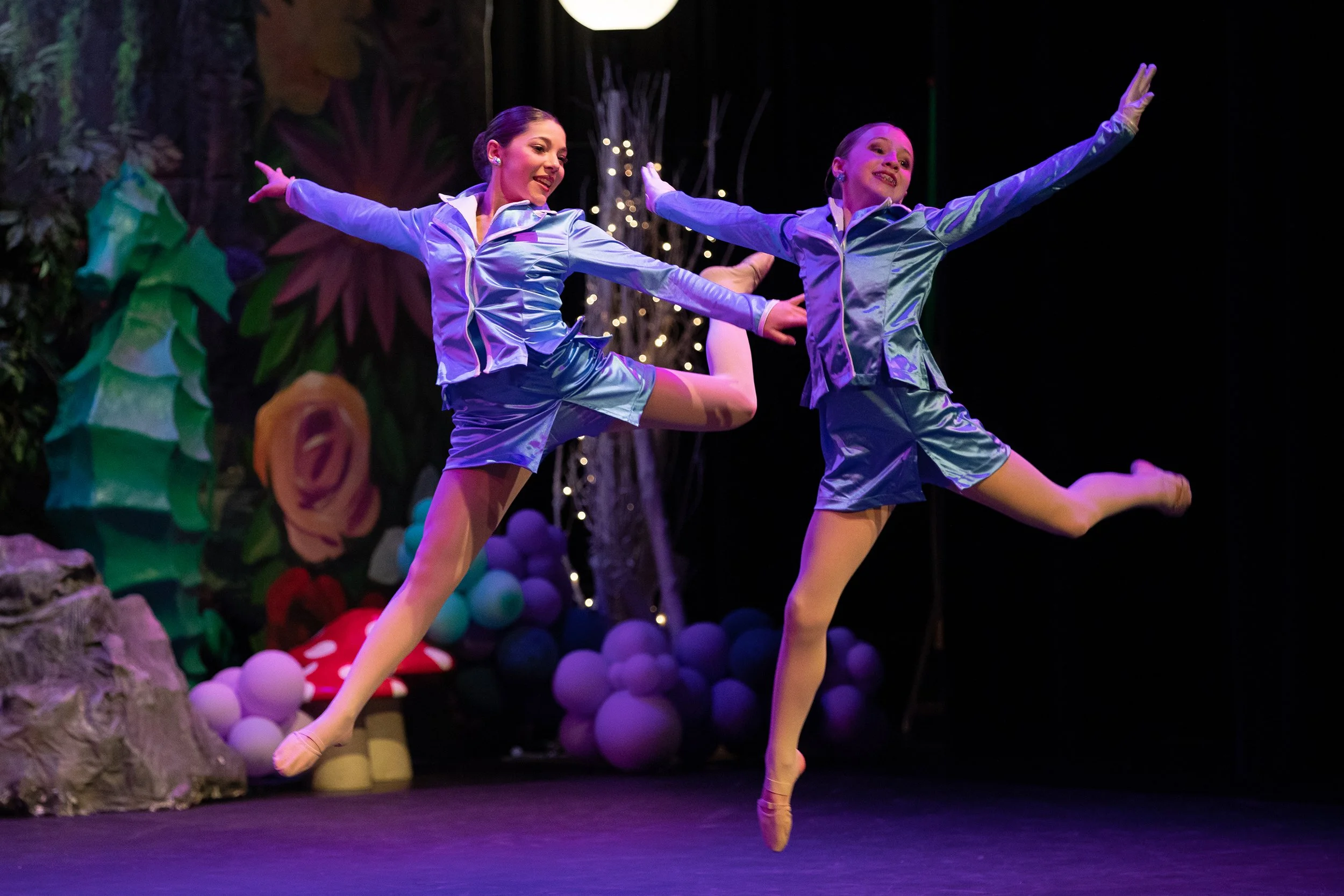 Two female dancers in shiny blue costumes performing ballet on stage with colorful whimsical background and props.