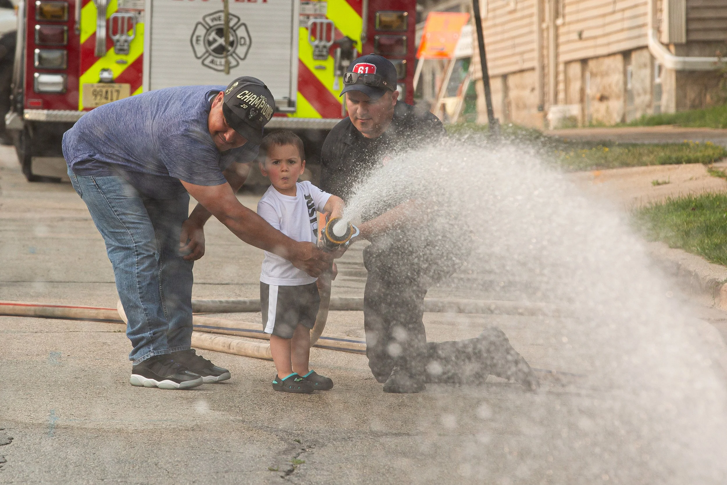A young boy stands in front of two firefighters during a fire safety demonstration, holding a fire hose with water spraying out. The firefighters are helping him aim the water, with a fire truck in the background.