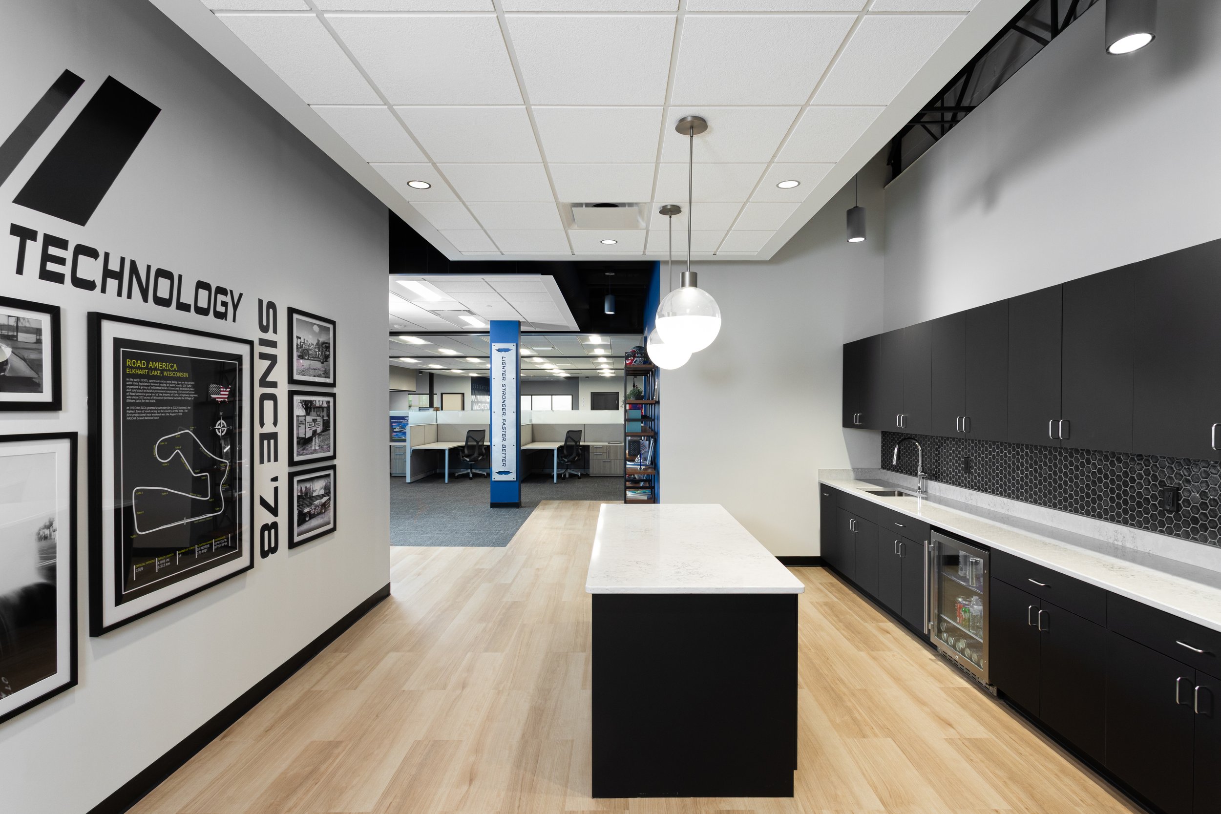 Modern office kitchenette with black cabinets, white countertop, and wooden floor. Decor includes framed posters and a wall with racing track map titled 'Technology Unite 78'.