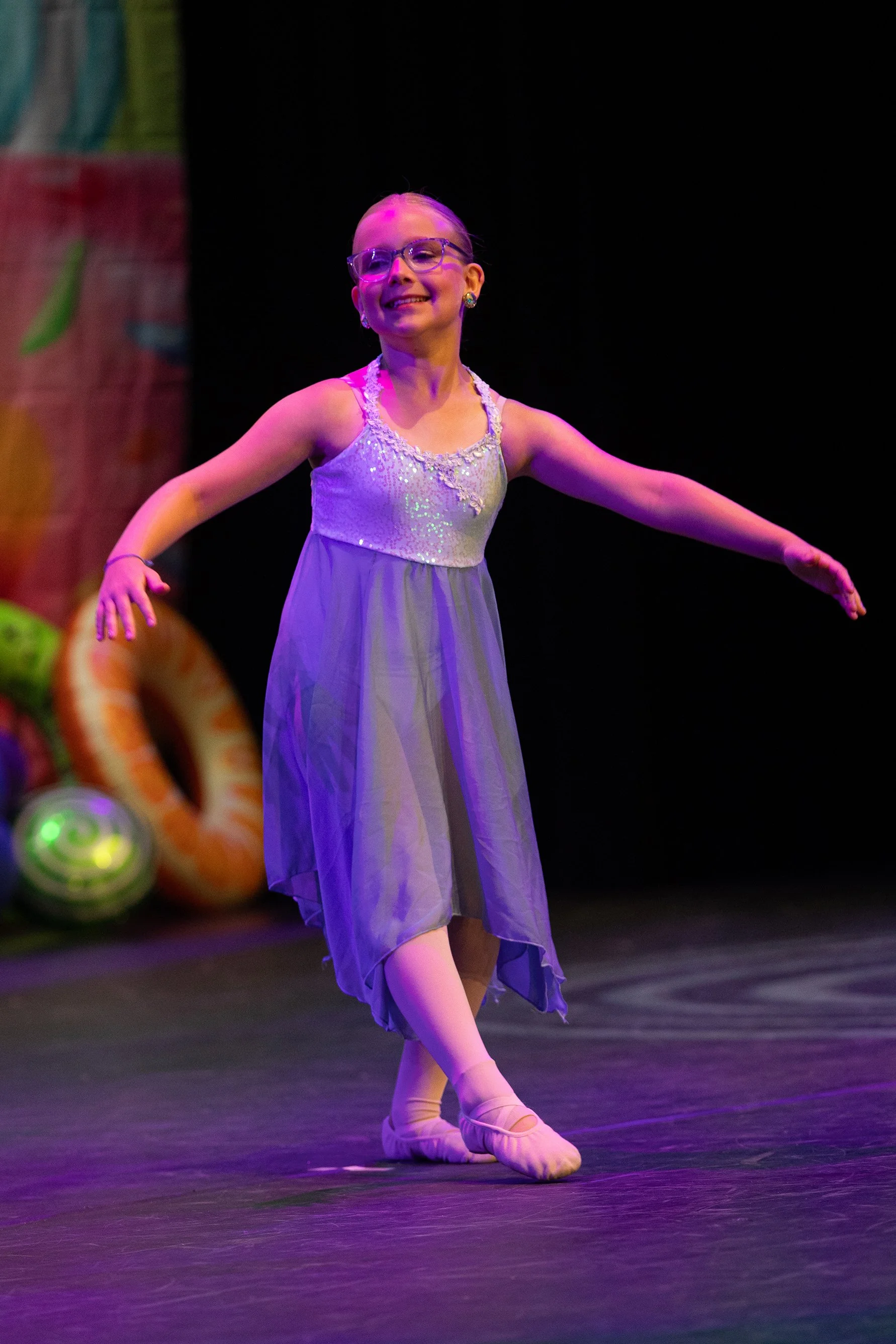 A young girl performing a ballet dance on stage, wearing a lavender dress and ballet slippers, smiling with arms outstretched in a graceful pose.