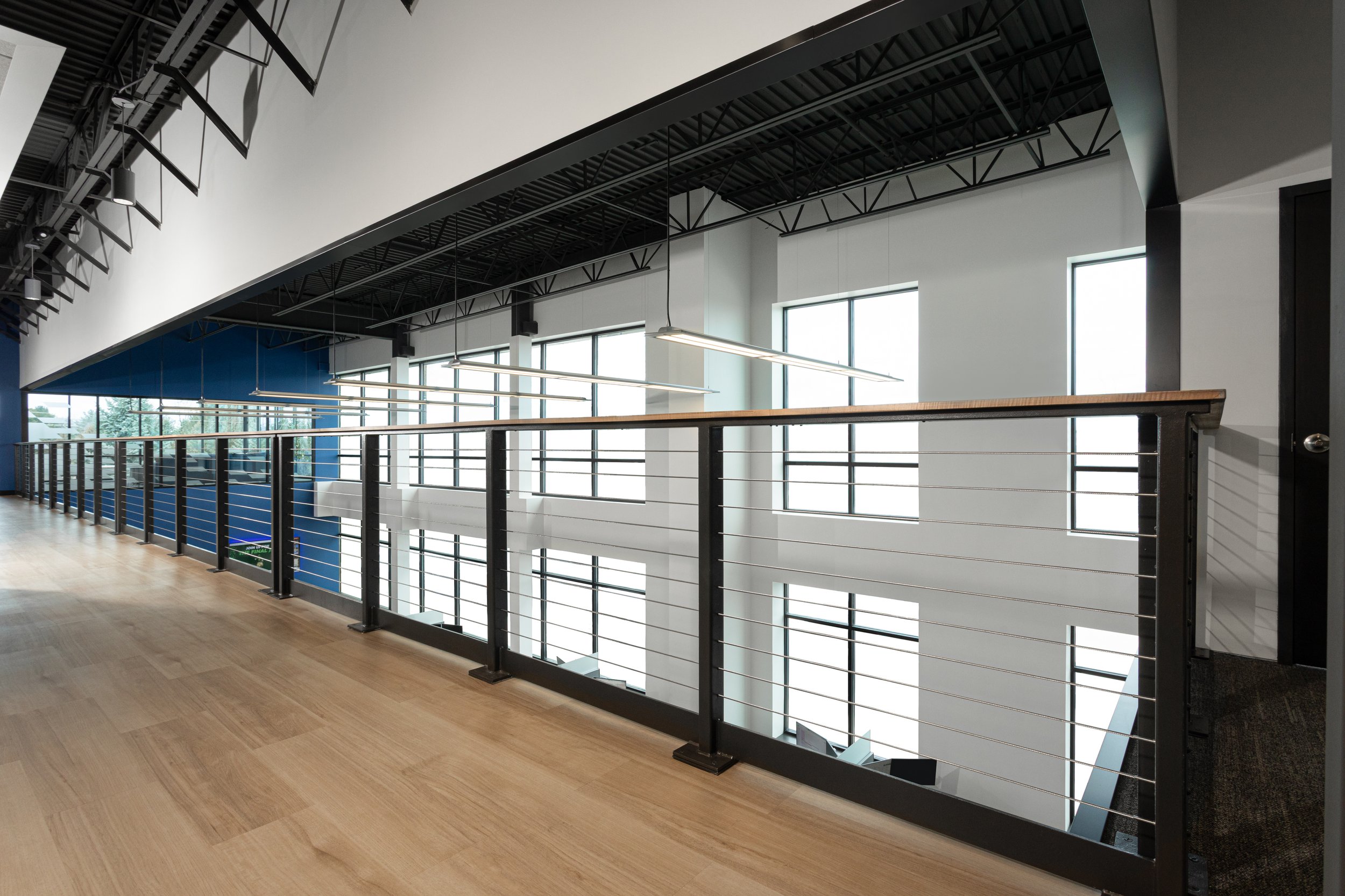 Modern interior corridor with large windows, wooden floor, metal railing, and hanging linear lighting fixtures in a commercial or office building.