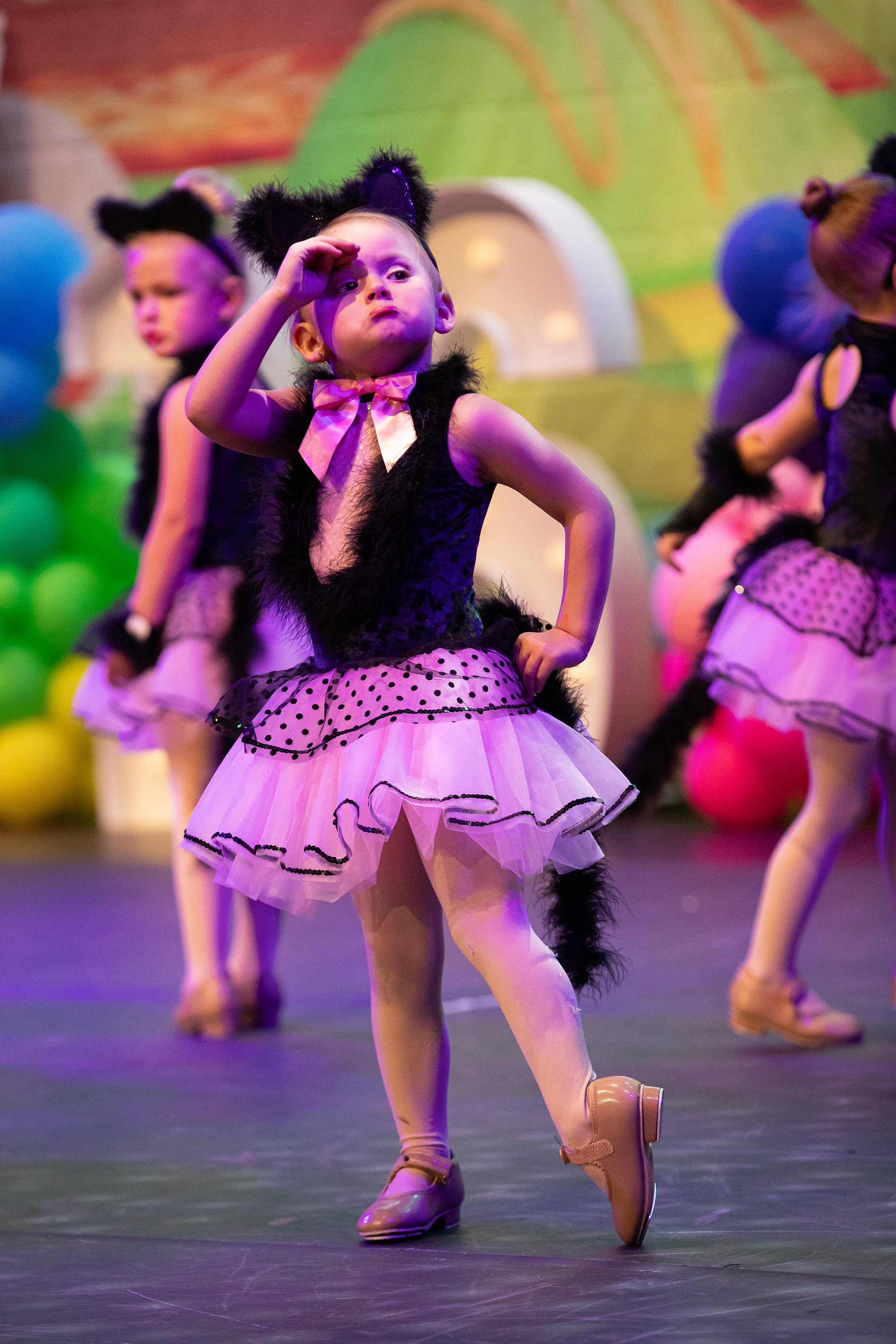 Young children dressed in black and pink cat costumes performing a dance on stage, with colorful balloons and a vibrant background.
