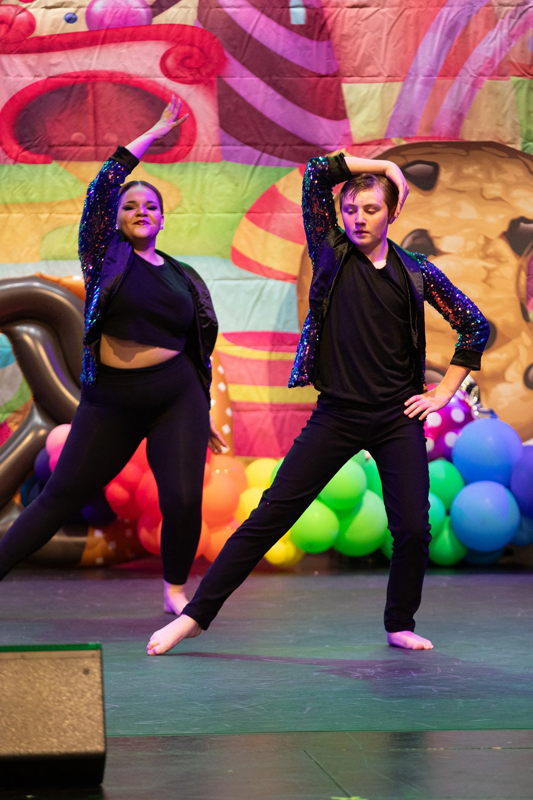 Two performers in black costumes with sequined jackets dancing on a stage with a colorful candy-themed backdrop and balloons.
