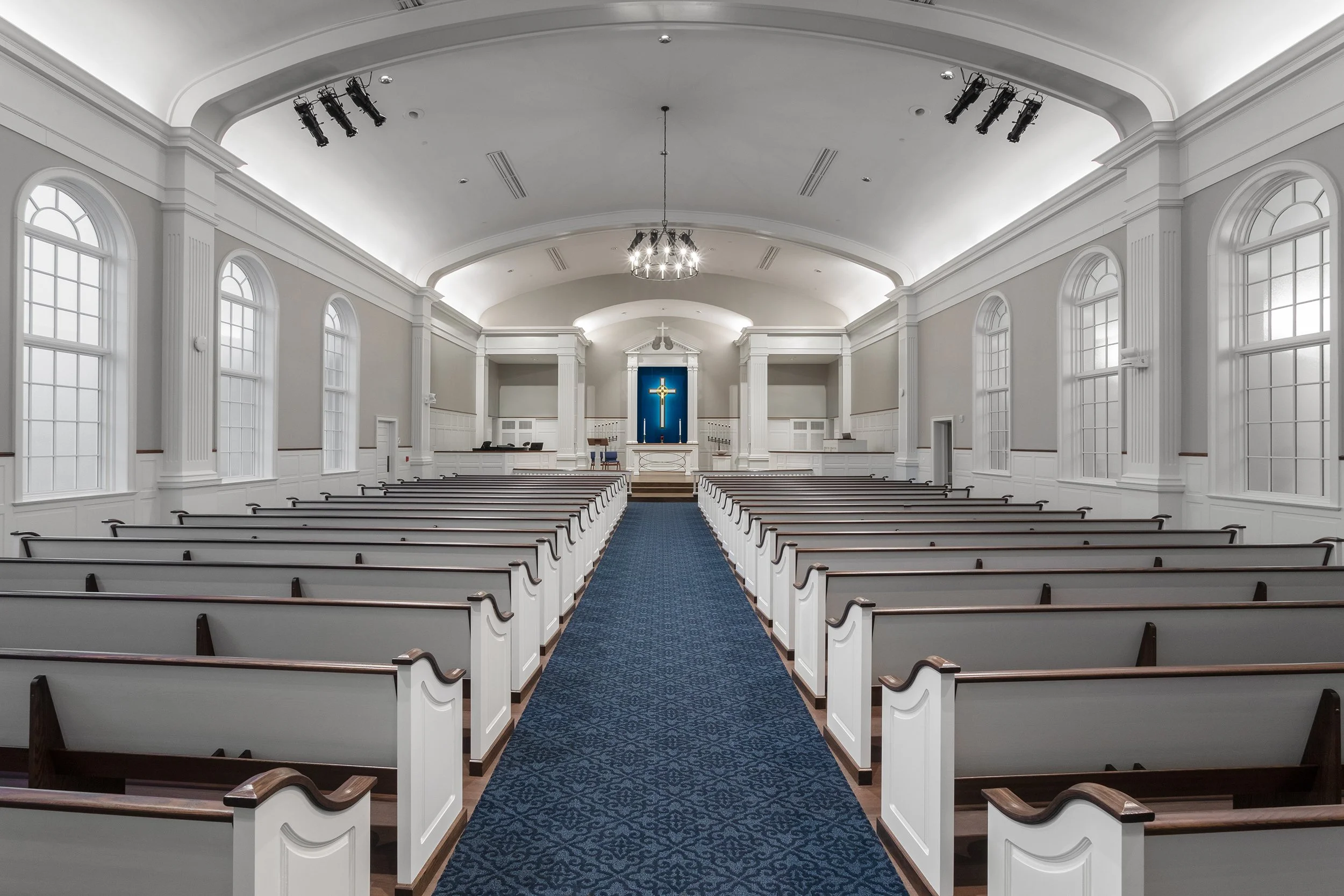 Interior of a church with white walls and large windows, rows of white pews, a blue carpet aisle, and a cross on a blue background behind the altar.