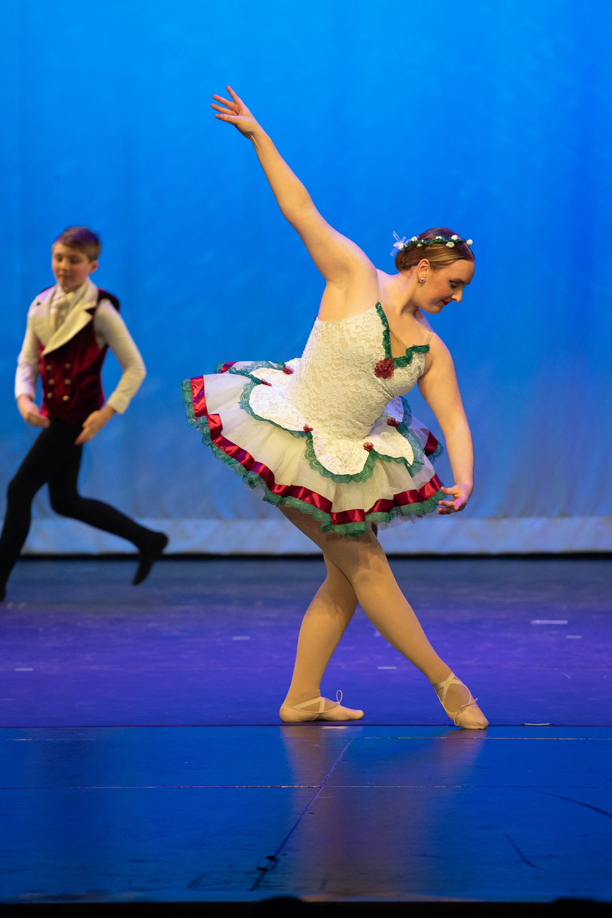 Ballet dancer in a floral-themed tutu performs on stage with a boy in a vest in the background.