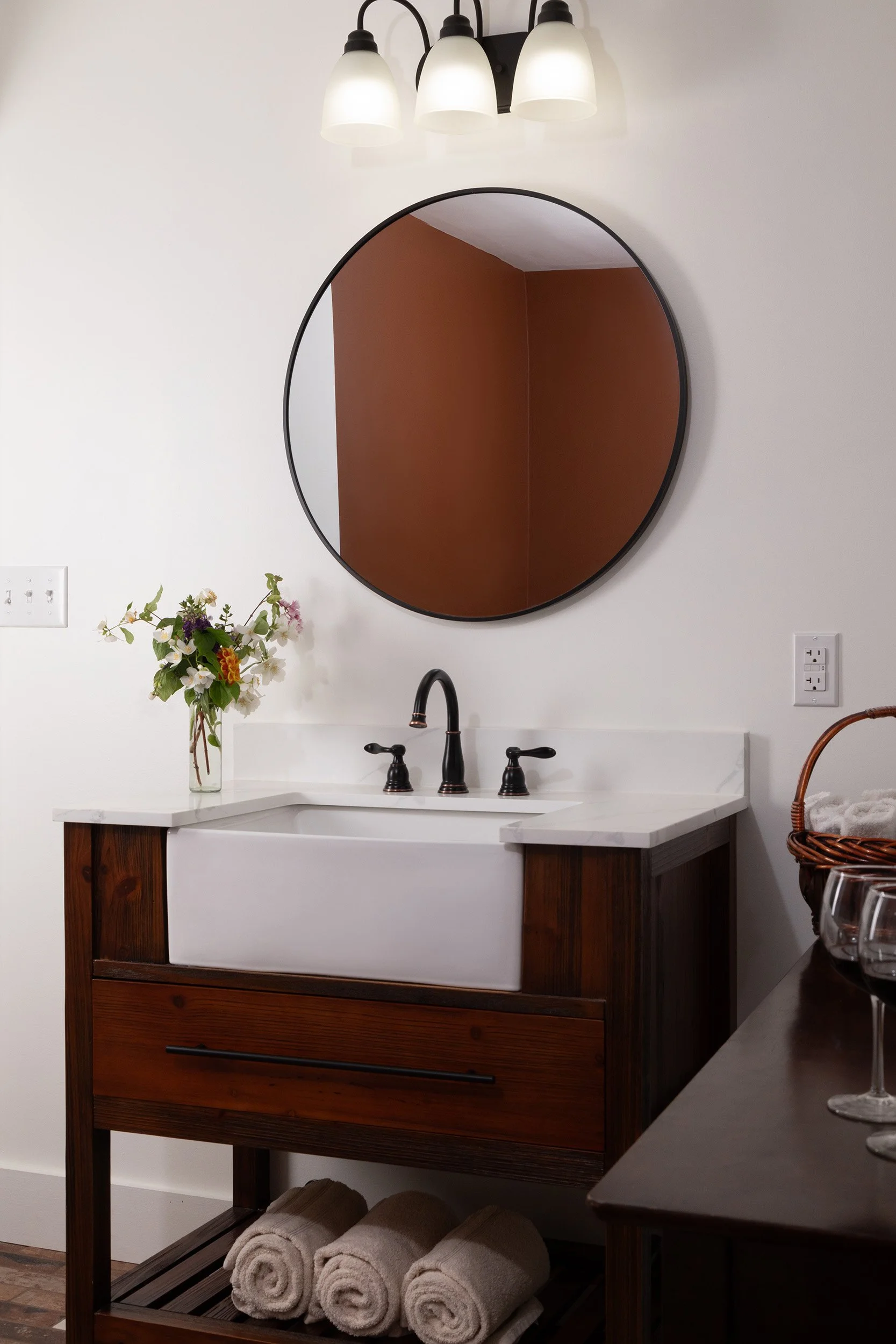 A bathroom vanity with a white farmhouse sink, a black faucet, and brown wooden accents. Above it, a round mirror with a black frame reflects a brown wall. To the left, a clear glass vase with colorful flowers sits on the counter, and below, rolled w
