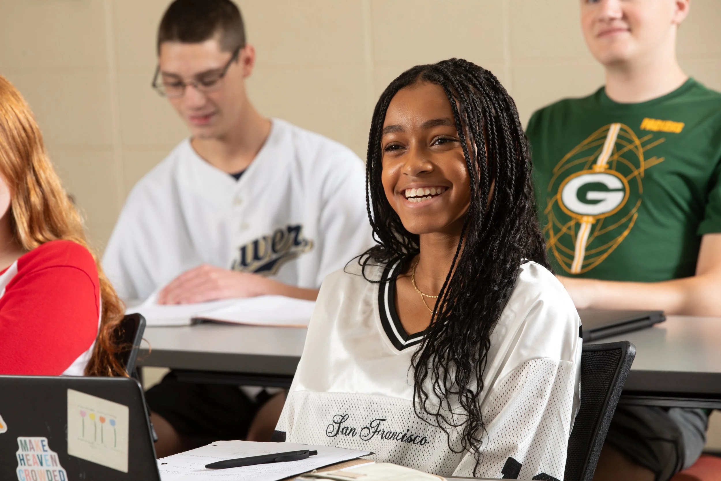A smiling African American girl with long braided hair sitting in a classroom, wearing a white and black shirt with 'San Francisco' written on it, surrounded by other students.