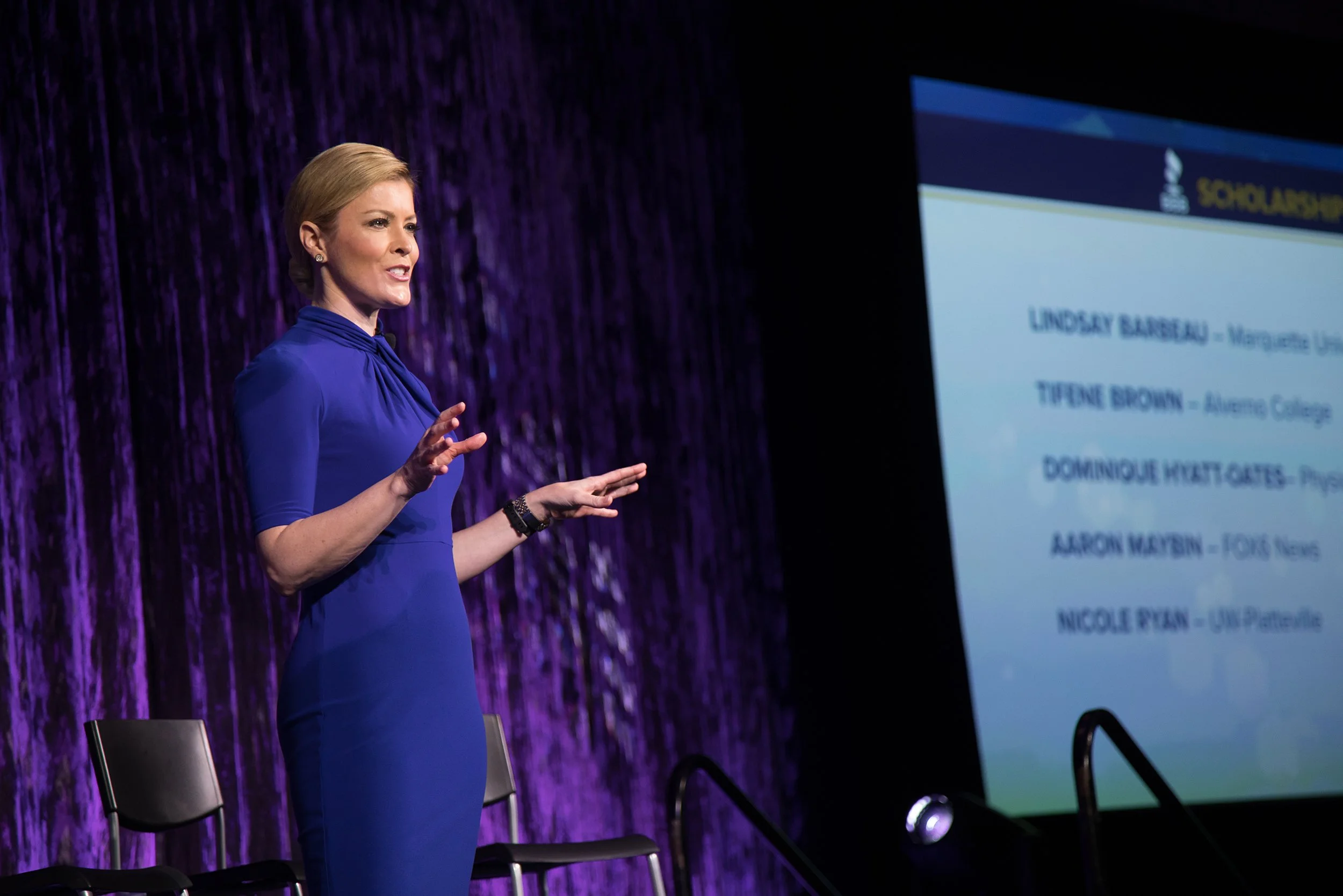 A woman with short blonde hair wearing a blue dress is giving a presentation on stage, with a large screen displaying her name, Lindsay Barbeau, and other partial text. The stage has a textured purple curtain background, and there are empty chairs in
