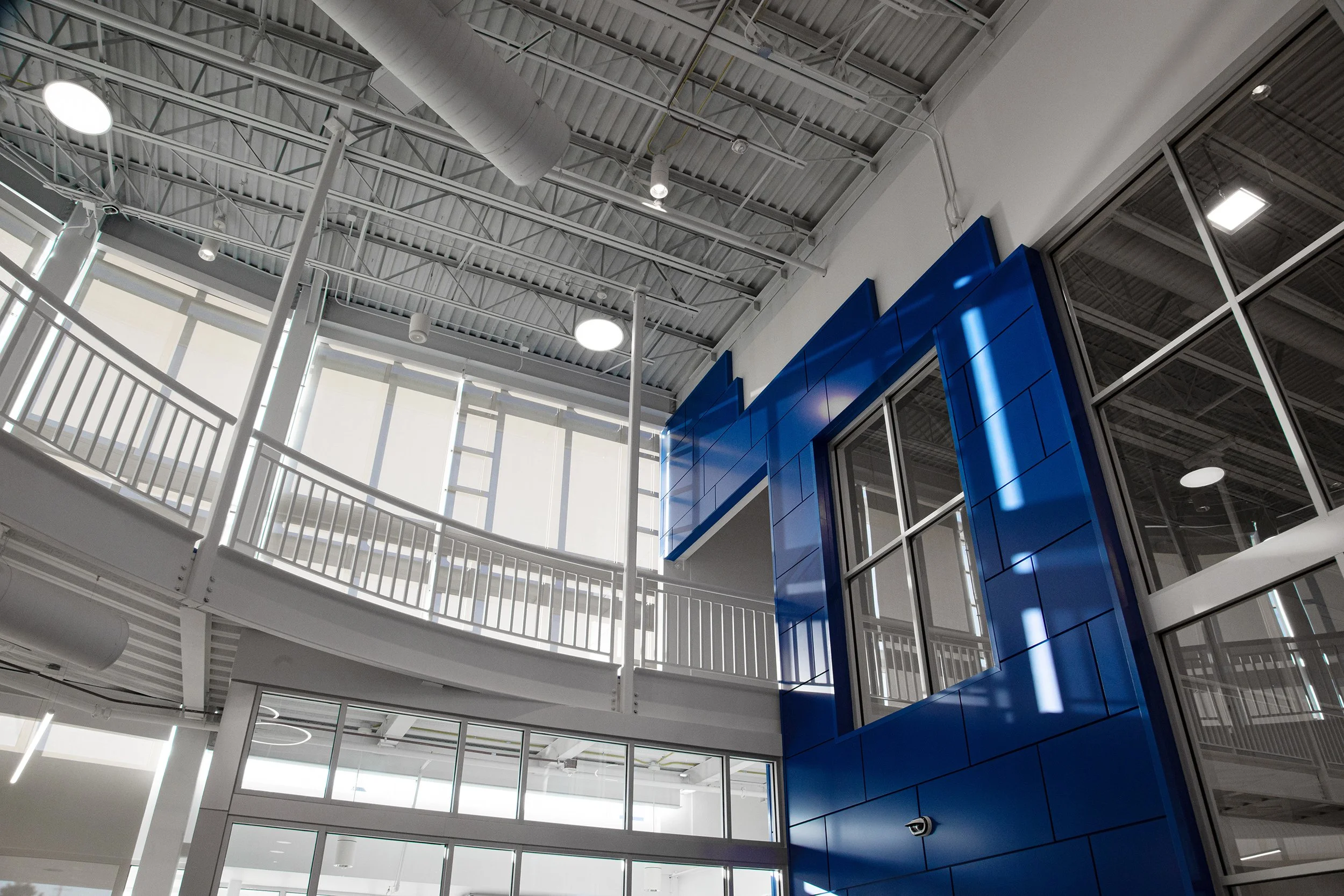 Interior view of a modern building with a blue wall, glass windows, metal balcony railing, and exposed industrial ceiling with lights.