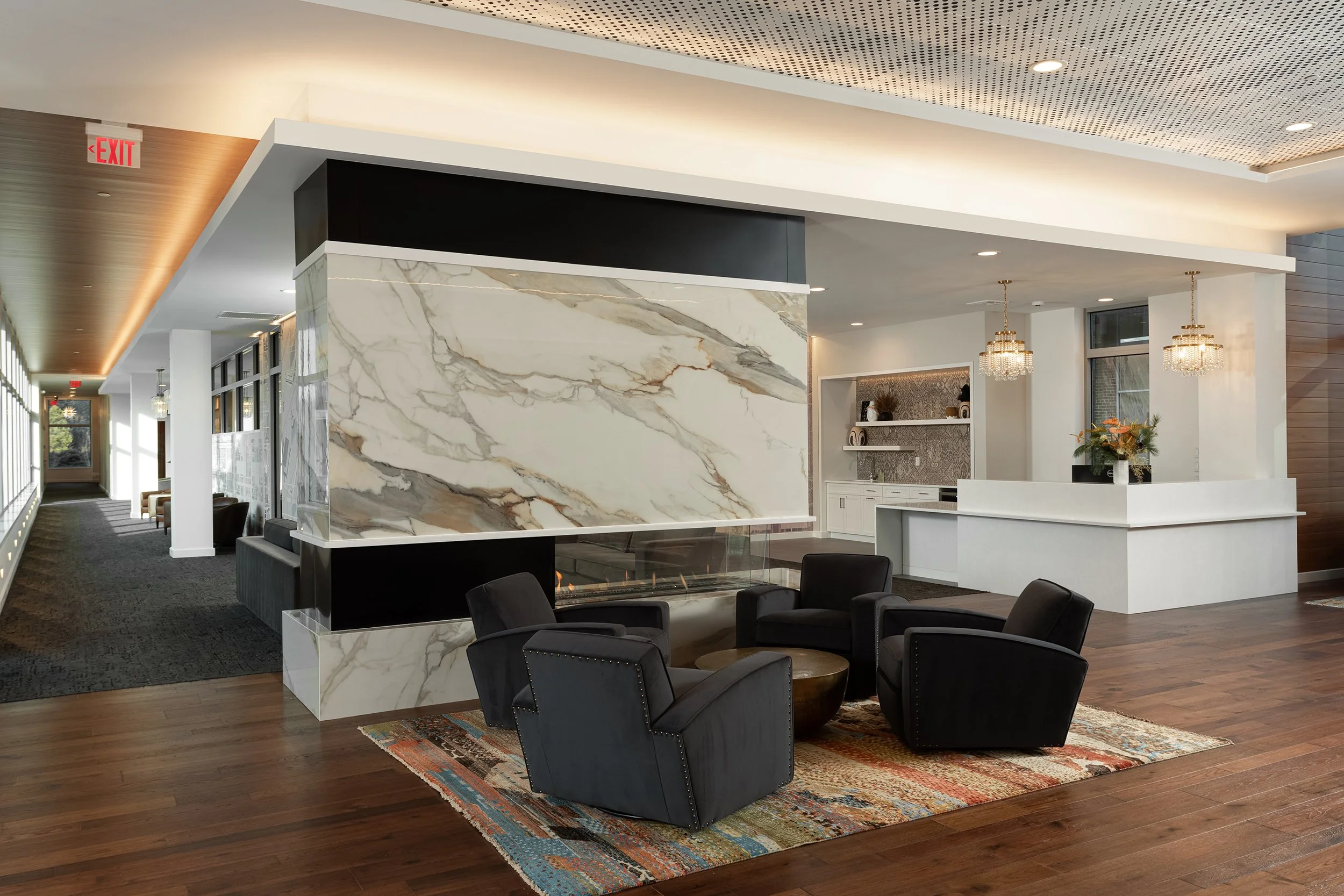 Modern hotel lobby with a large marble fireplace, black leather armchairs, a colorful rug, chandeliers, and a reception desk, with windows along the hallway.