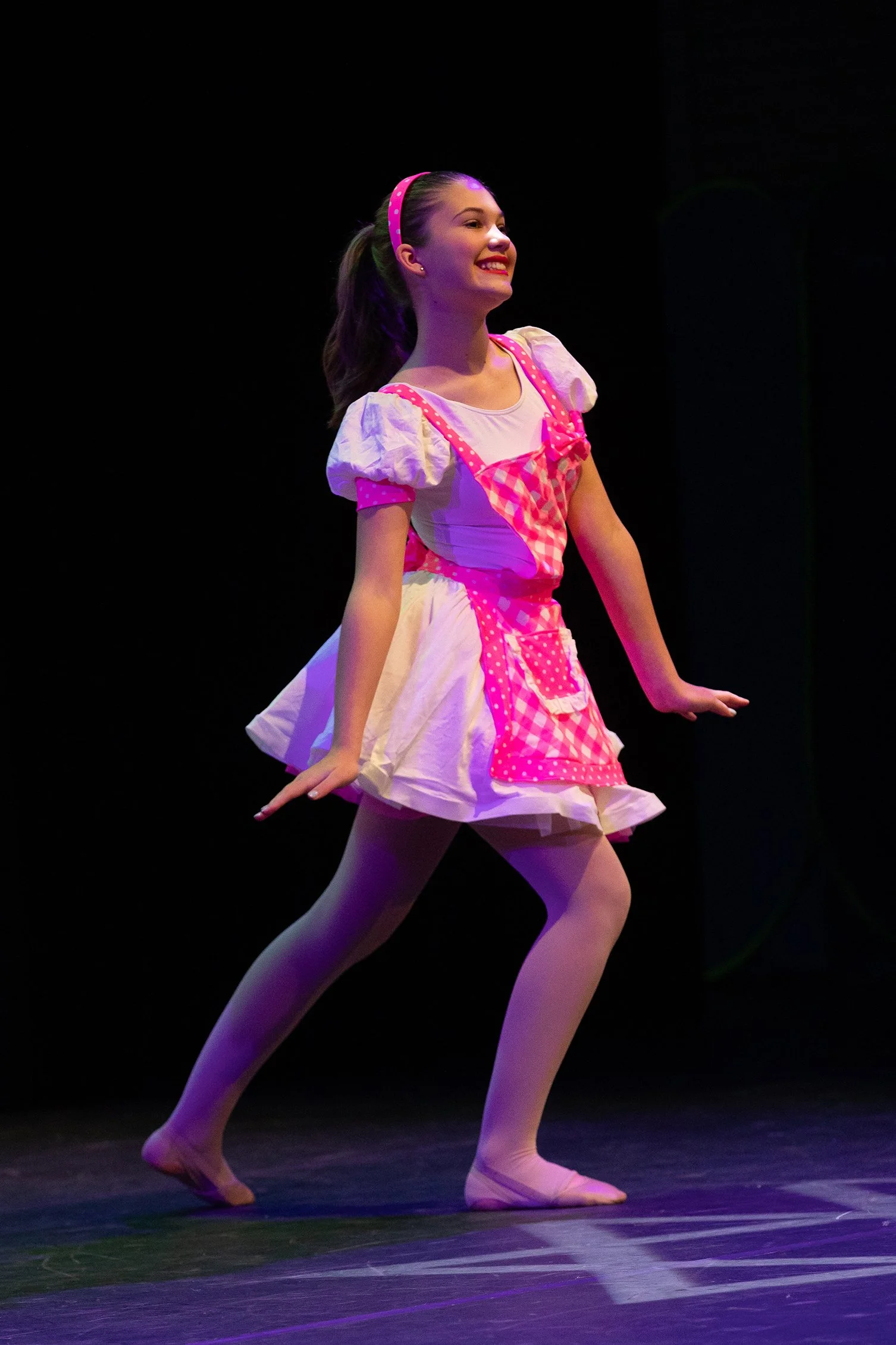 A young girl dancing on stage wearing a pink and white costume with a checkered apron and pink headband, smiling.