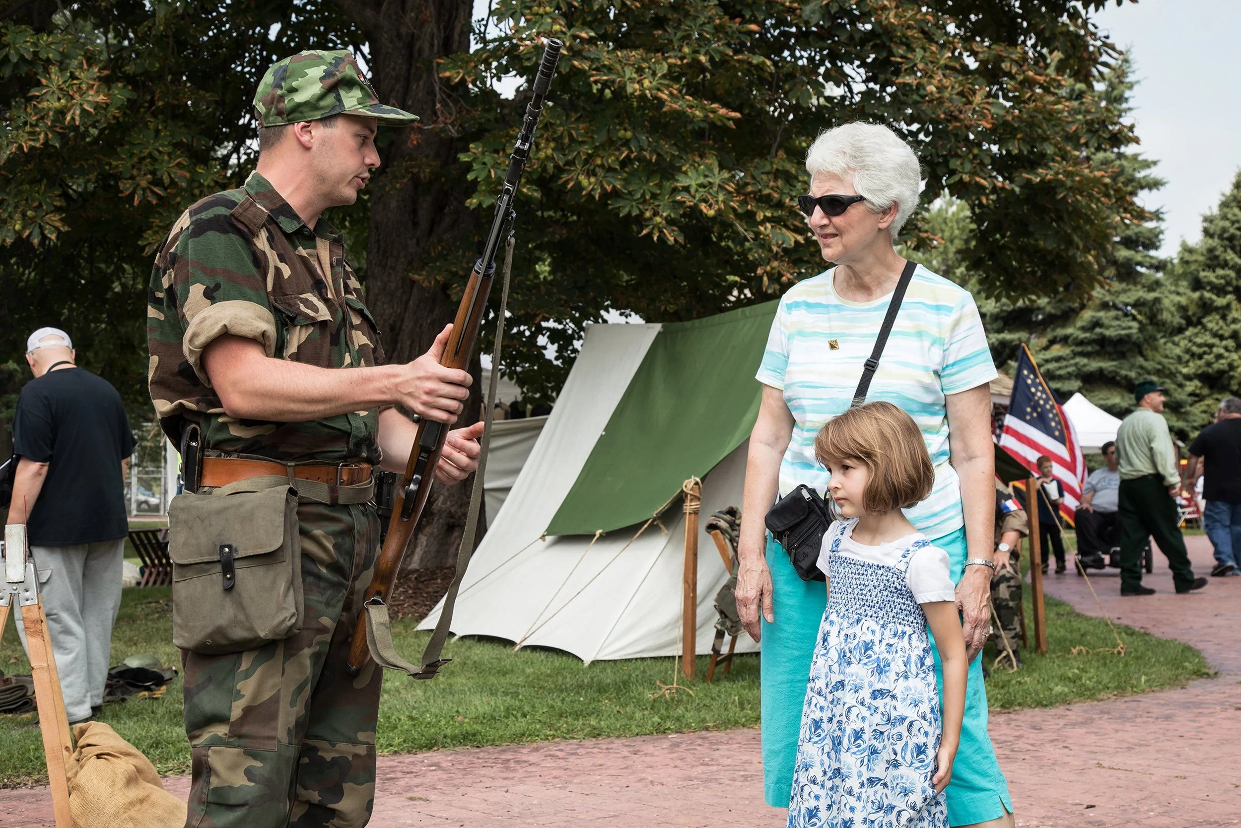A man in military uniform holding a rifle. An elderly woman wearing sunglasses and a striped shirt, standing with two young girls, are talking to the man at an outdoor event with tents and American flags visible in the background.