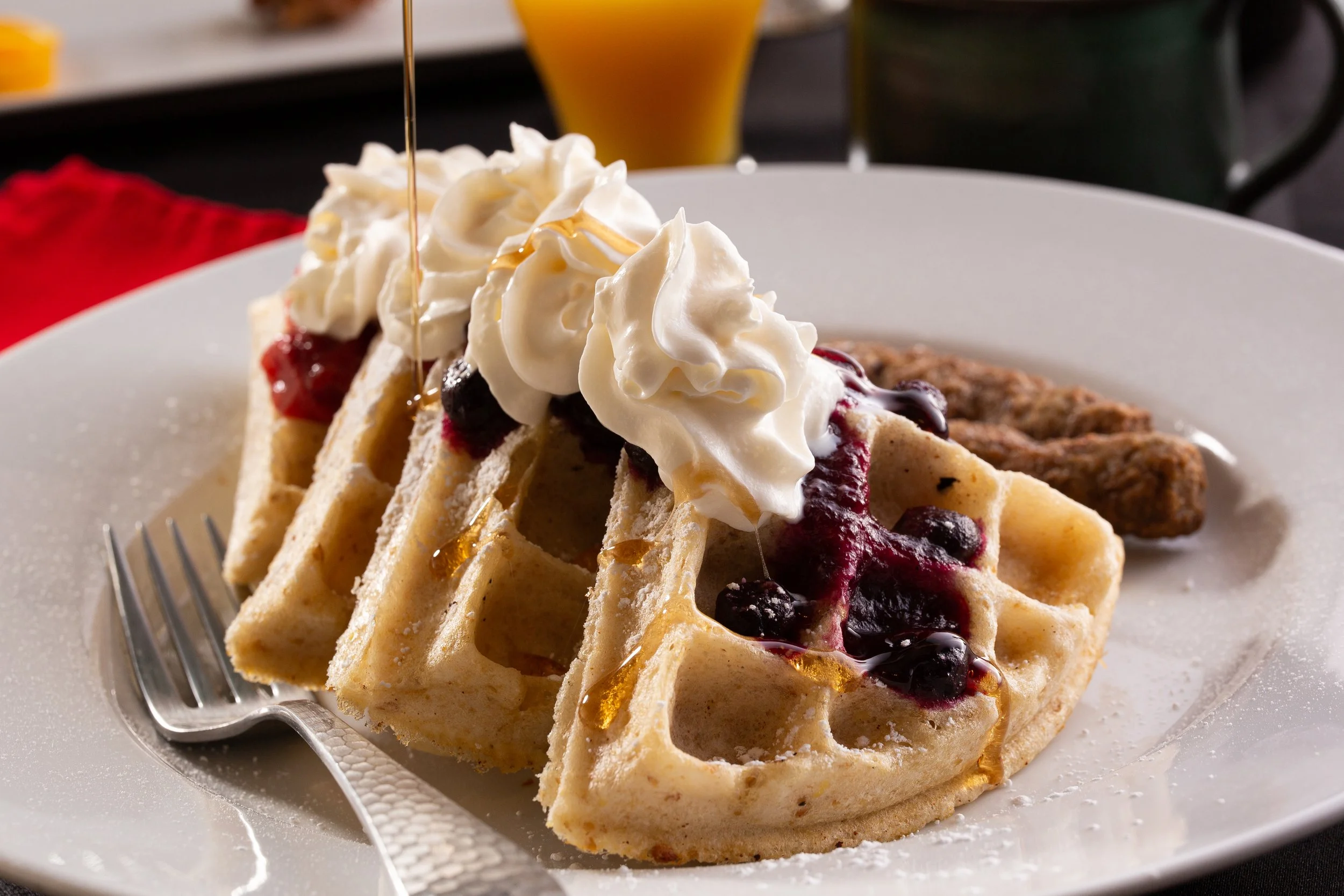 A plate of waffles topped with whipped cream, berries, and syrup, with bacon in the background.