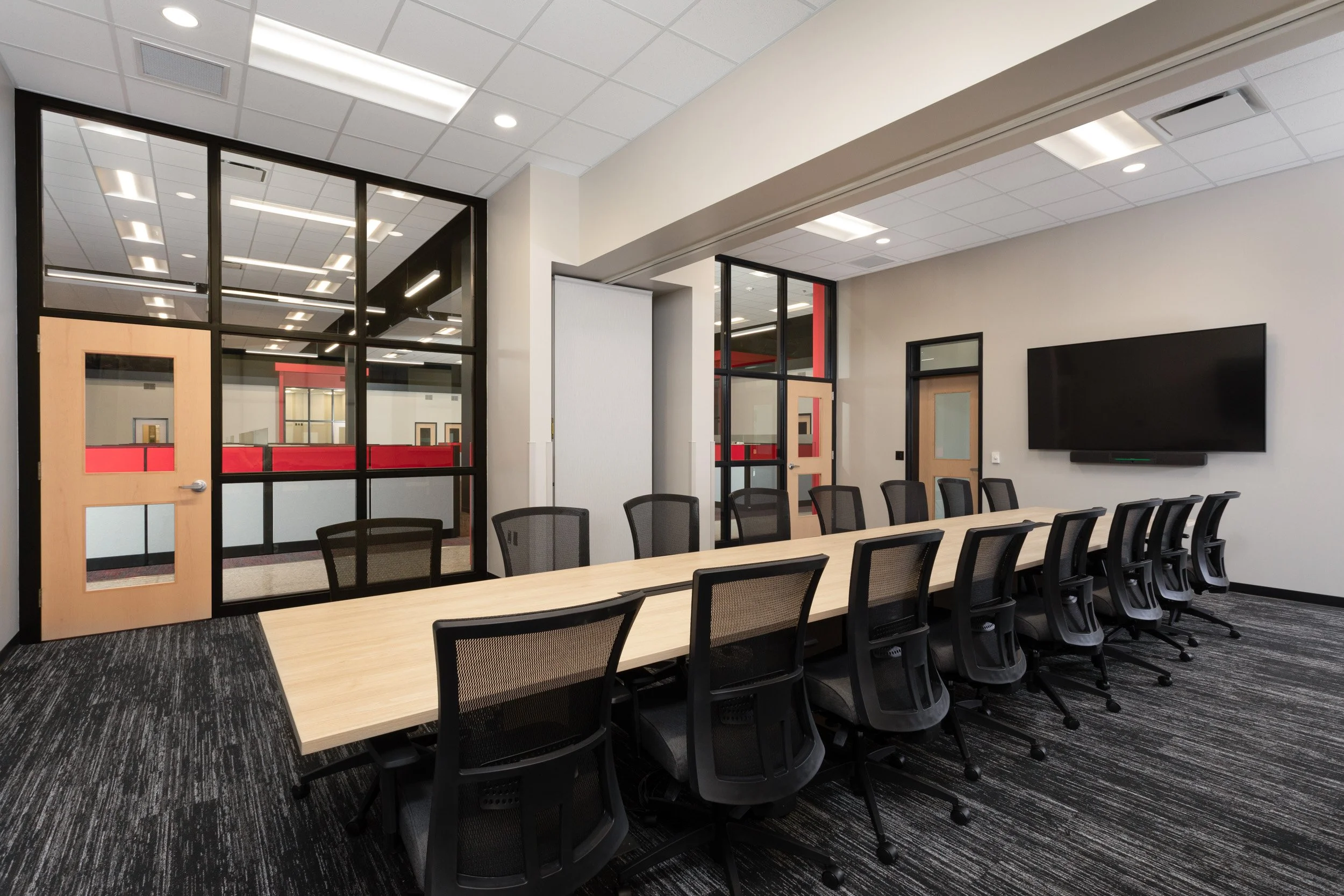 Conference room with a long table, black mesh chairs, a large wall-mounted TV, glass walls with black frames, and a entry door with windows.