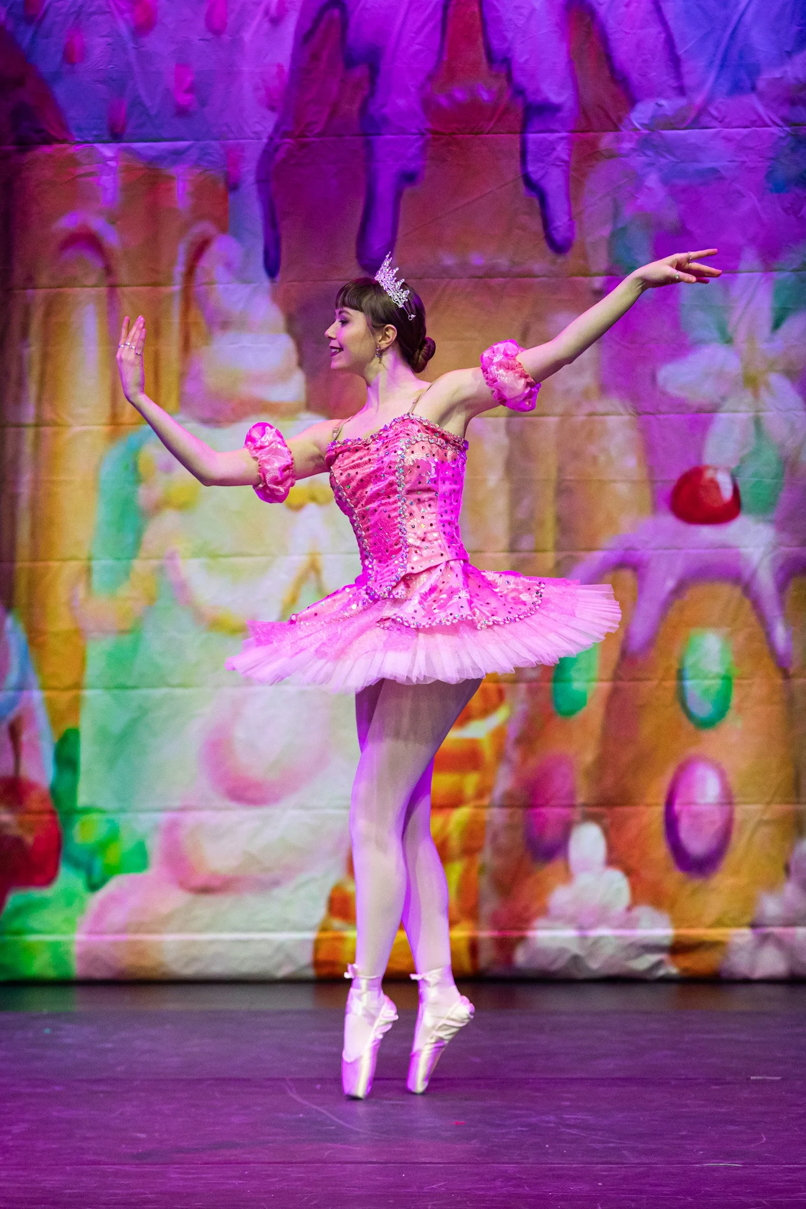 Ballet dancer in a pink tutu and crown performing on stage with a colorful background.