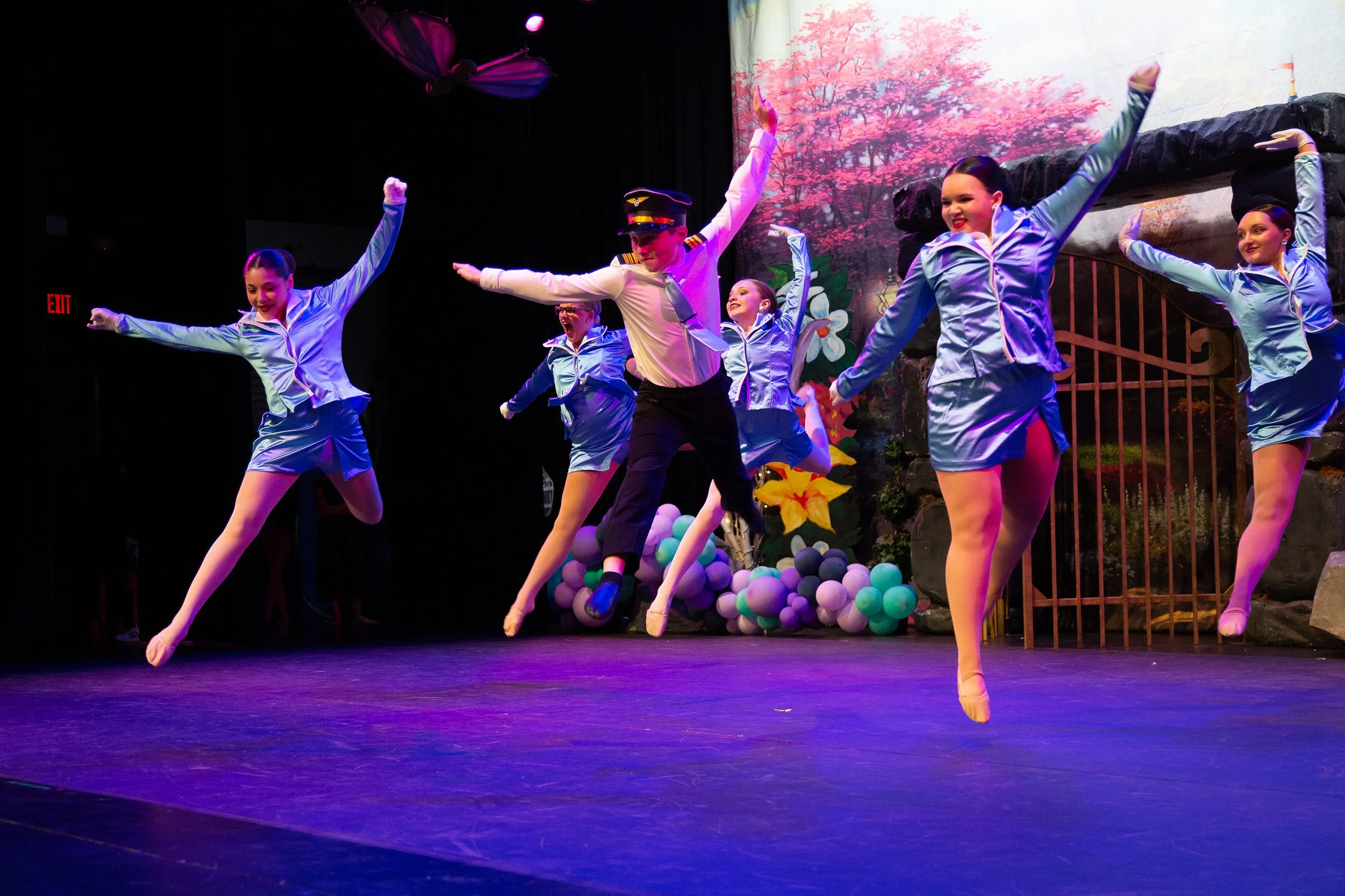 Group of ballet dancers on stage performing a leap, with a scenic backdrop of flowers and trees, and decorated with balloons.