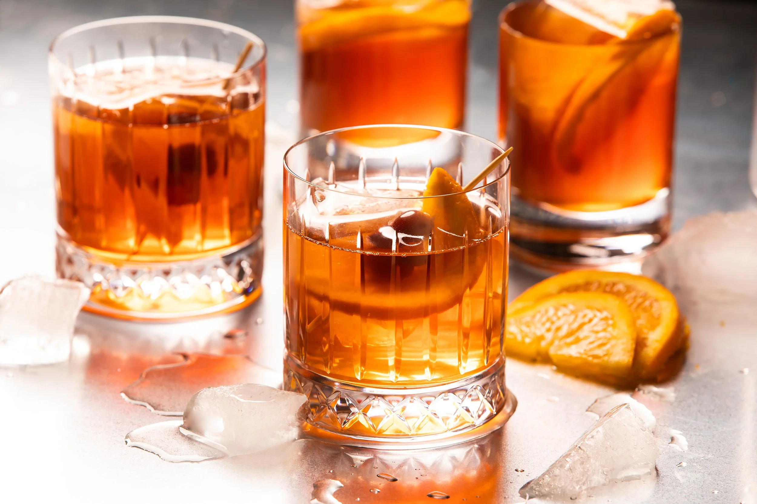 Four glasses of iced orange cocktails with lemon garnishes, ice cubes, and orange slices on a metal surface. Photo by product photographer Tom Krueger, Milwaukee, Wisconsin.