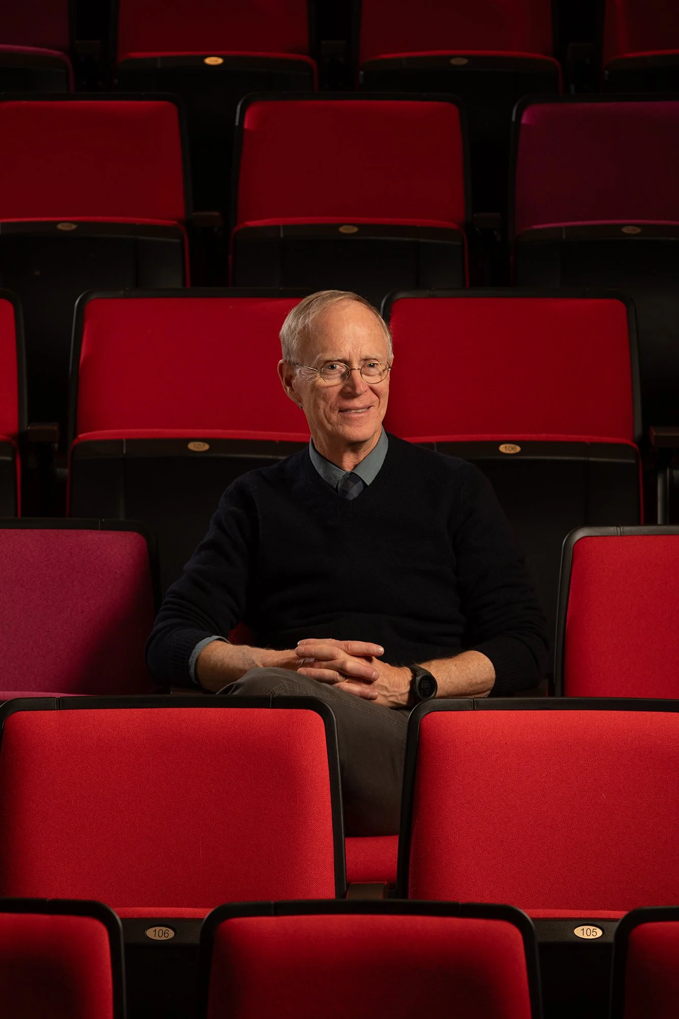 An older man with glasses, wearing a dark sweater, sitting in an empty theater with red seats, smiling and looking to the side.