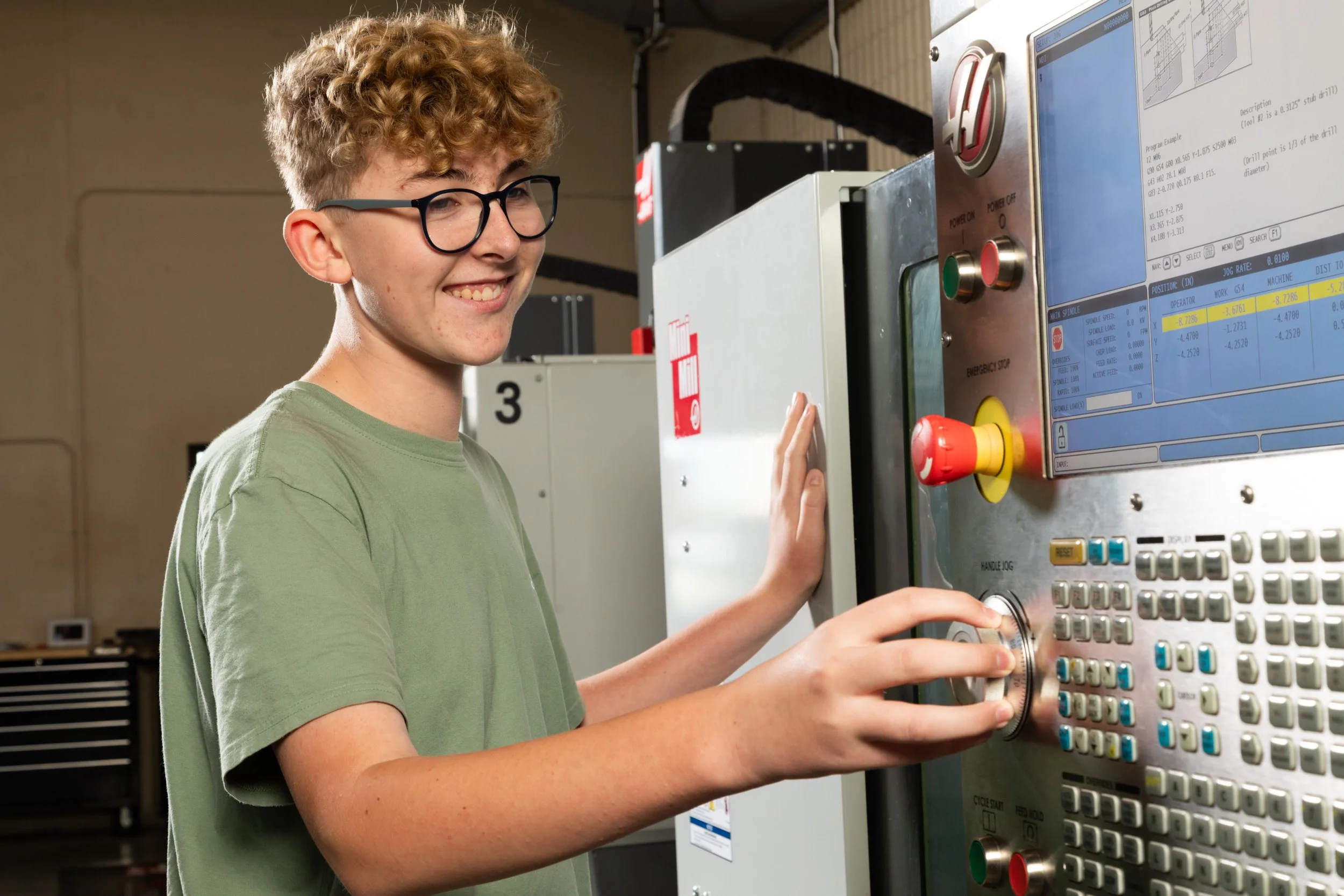 A young person with curly blond hair and glasses operating a CNC machine in a workshop, smiling while pressing buttons on the control panel.