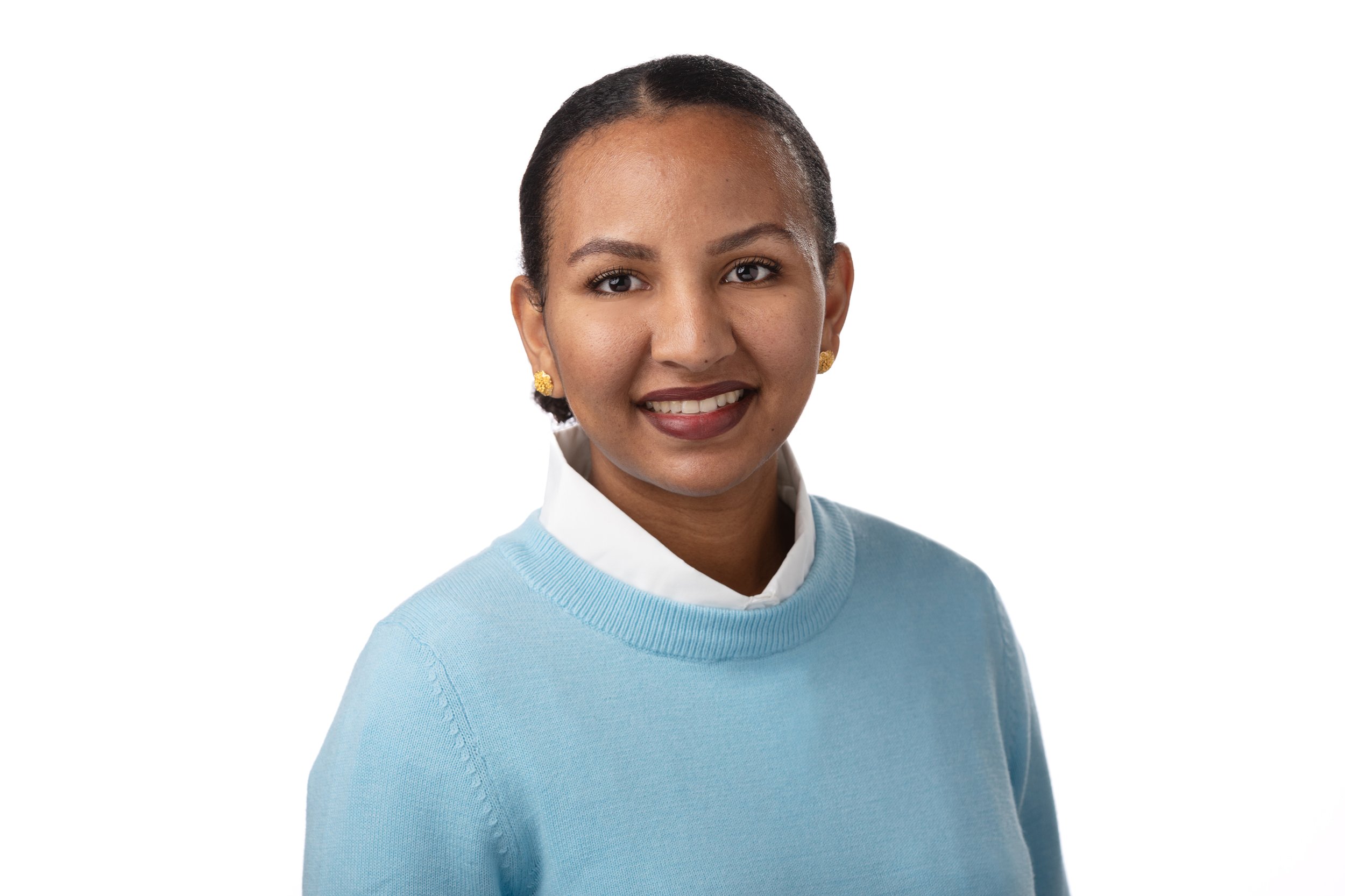 A woman smiling, wearing a light blue sweater and a white collared shirt with gold earrings, against a white background.