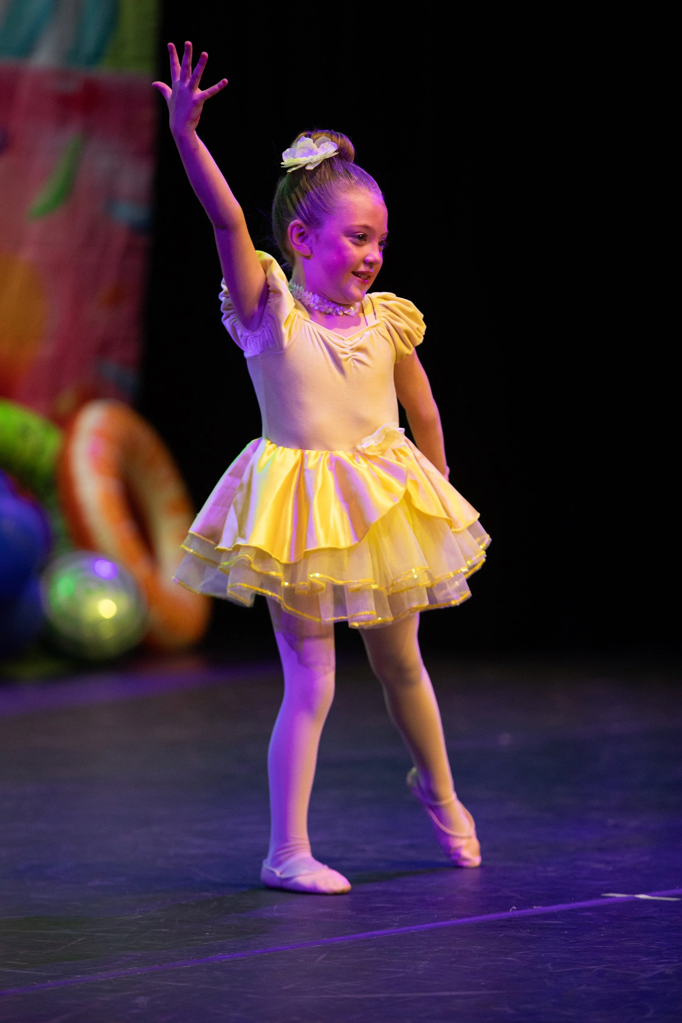 Young girl in a yellow dress performing ballet on stage with colorful balloon decorations in the background.