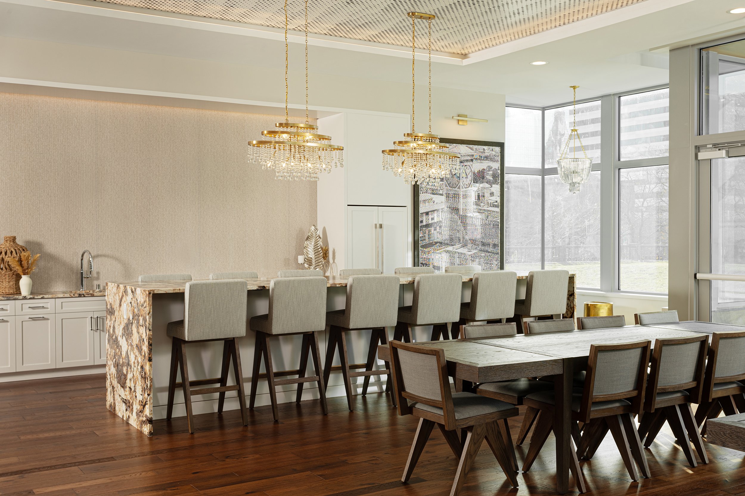 Modern dining area with a long table, beige upholstered chairs, and decorative chandeliers hanging from a ceiling with recessed lighting. Large windows provide natural light.