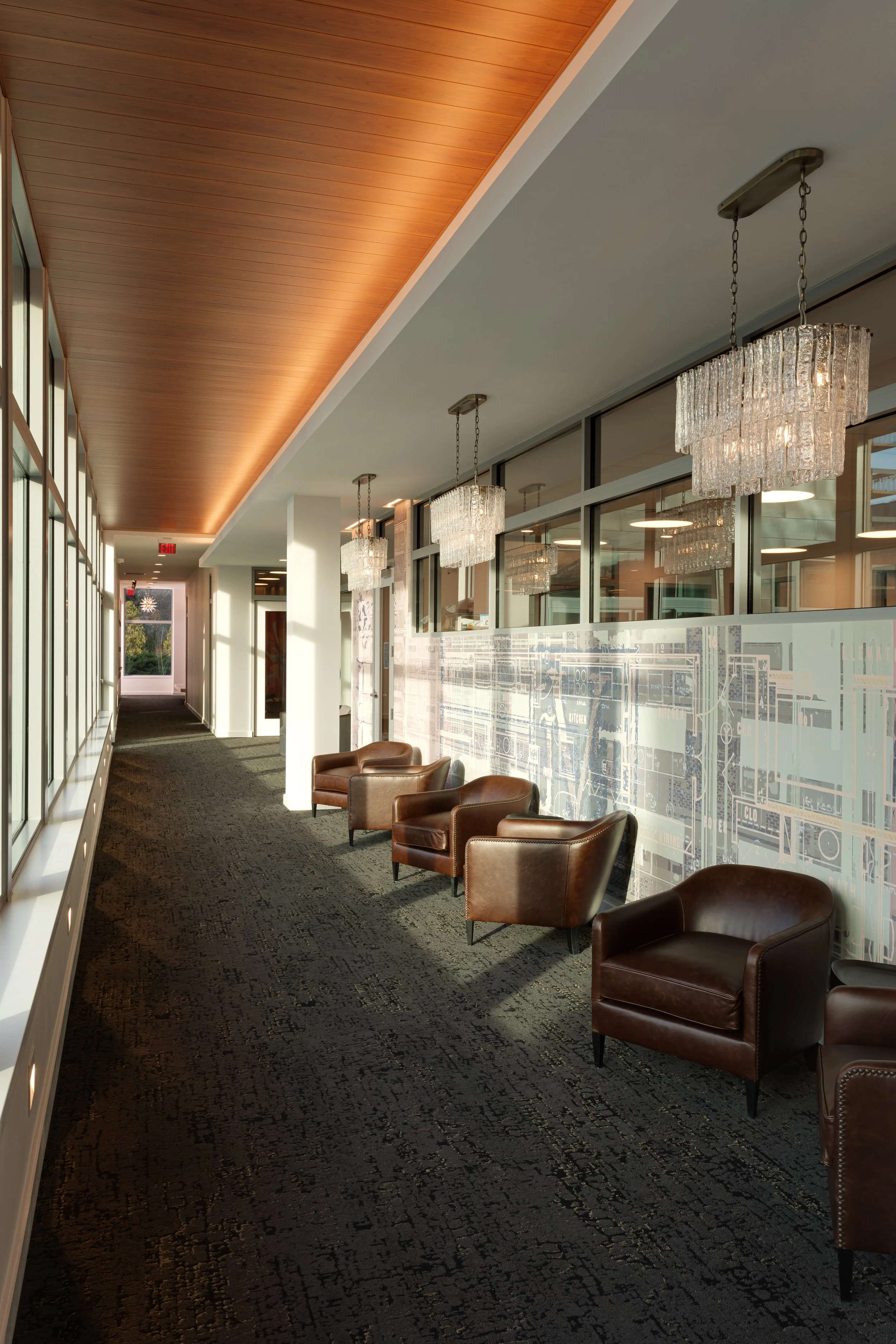 Modern office hallway with large windows, leather chairs, and elegant chandeliers.