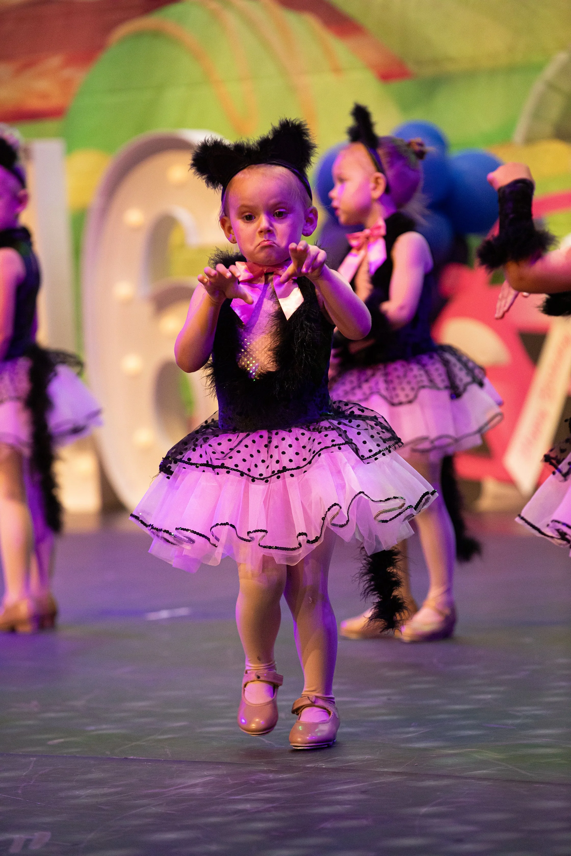 Young girl dressed as a black cat in a pink and black tutu, performing a dance on stage with other children in similar costumes.