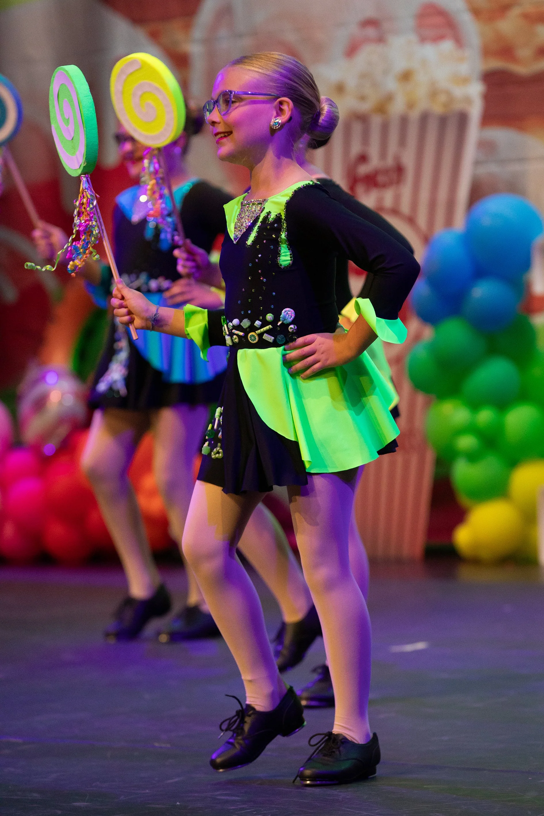 A young girl with blonde hair tied in a bun, wearing glasses and a colorful costume, is dancing on stage holding a lollipop-shaped prop. Other children in similar costumes are in the background, and colorful balloons decorate the stage.